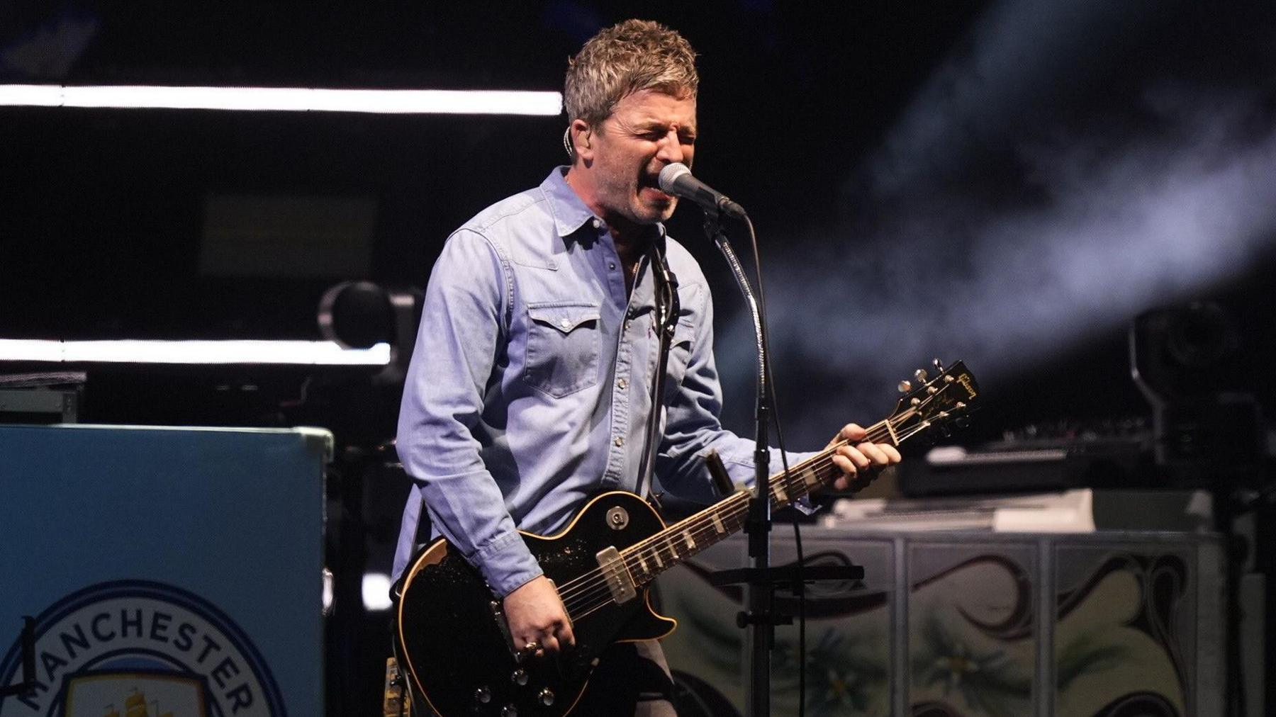 Noel Gallagher in denim shirt singing and playing a black Gibson Les Paul guitar on stage in Cardiff.