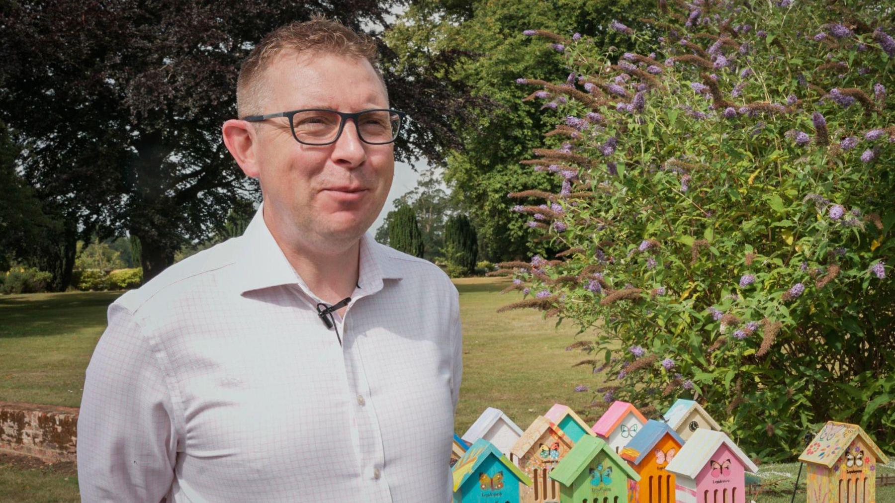 Leighton Reid with short hear wearing glasses and a white shirt looks at the camera with the greenery of a garden behind him and brightly coloured miniature houses designed for butterflies shown on the ground.