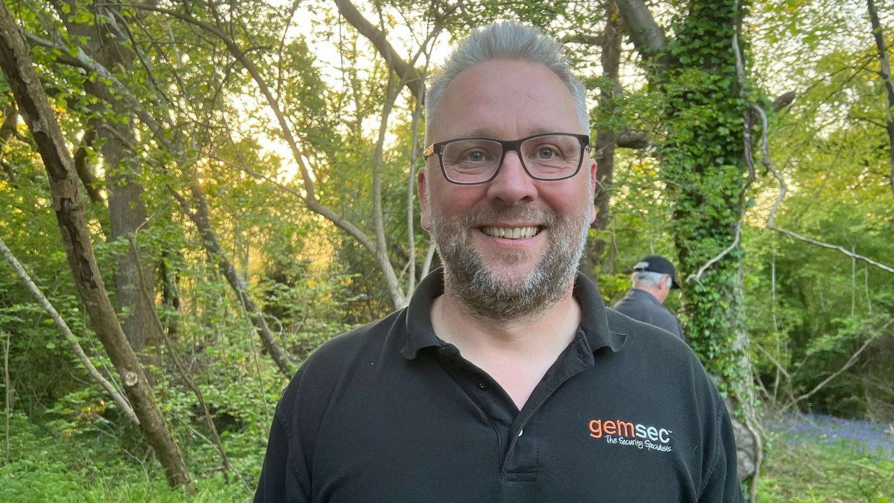 Mr Winterson has grey hair, a grey beard, and wears a black branded polo and black square glasses. He is smiling at the camera in woodland, and there are trees and bluebells in the background.