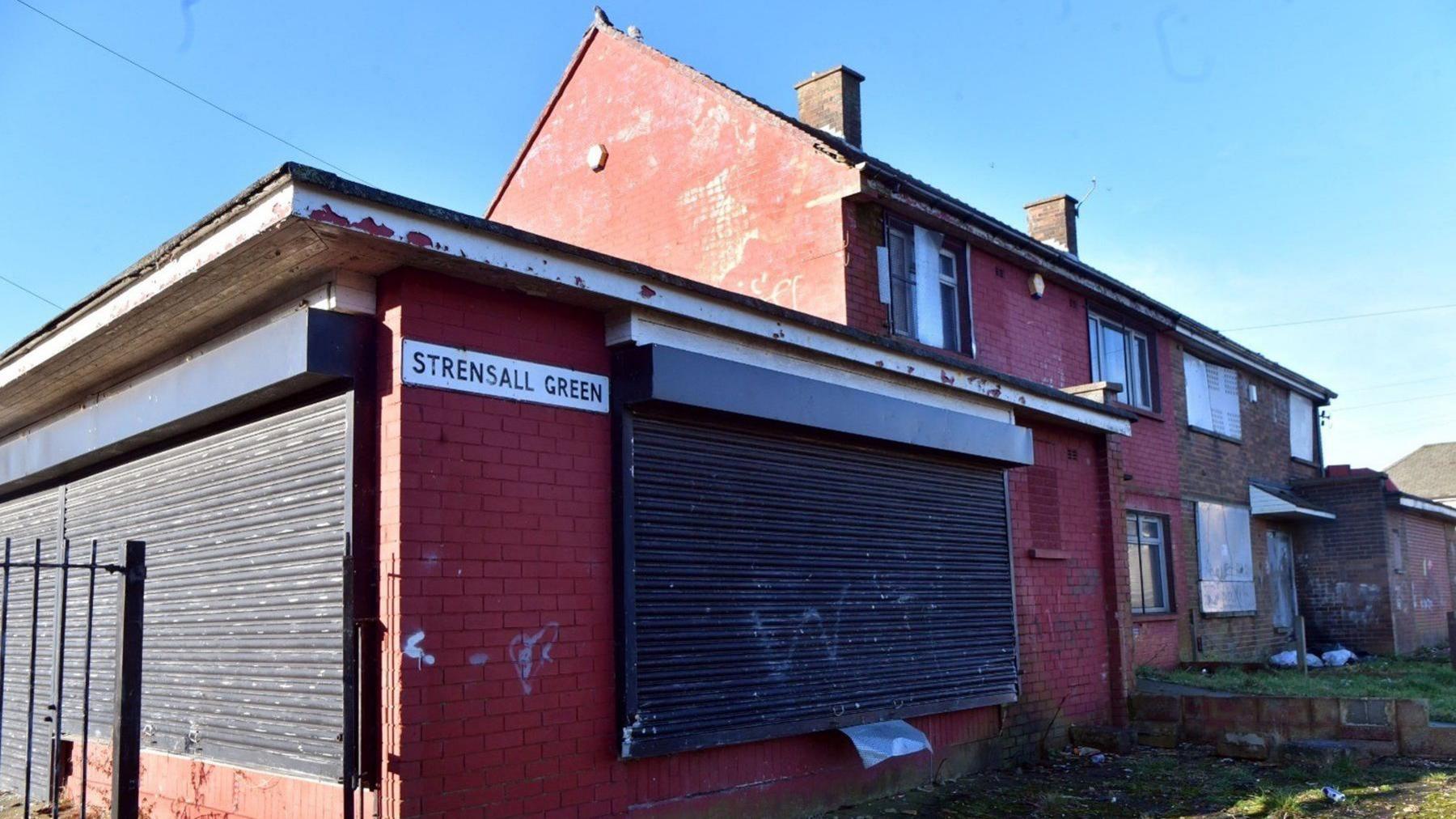 Dilapidated properties, one with a metal screen over its window with a road sign saying "Strensall Green"