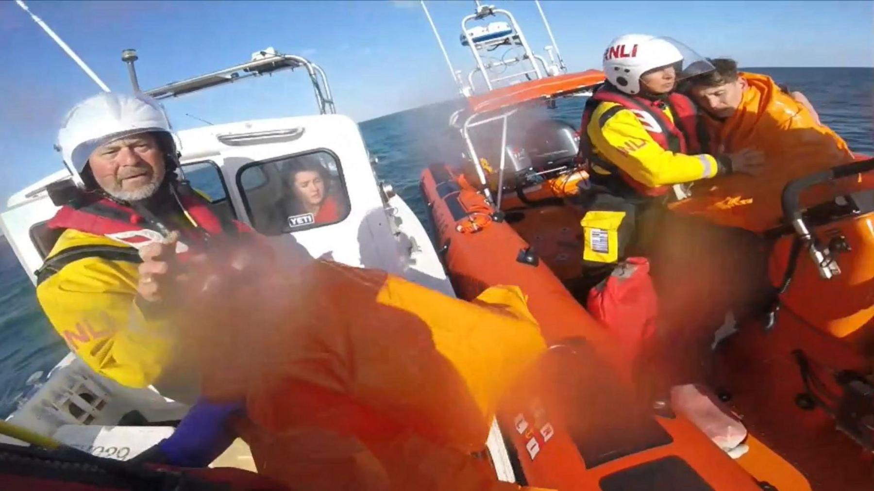 A still from the body warn camera of an RNLI rescuer showing the orange RIB lifeboat alongside the small white fishing vessel. In the RIB, a lifeboat volunteer has his arm around Sebastian Pixley who is wrapped in a plastic sheet and his eyes are closed. Another volunteer is standing on the deck of the fishing vessel and Kyra Farnum can be seen through the window of the cabin.