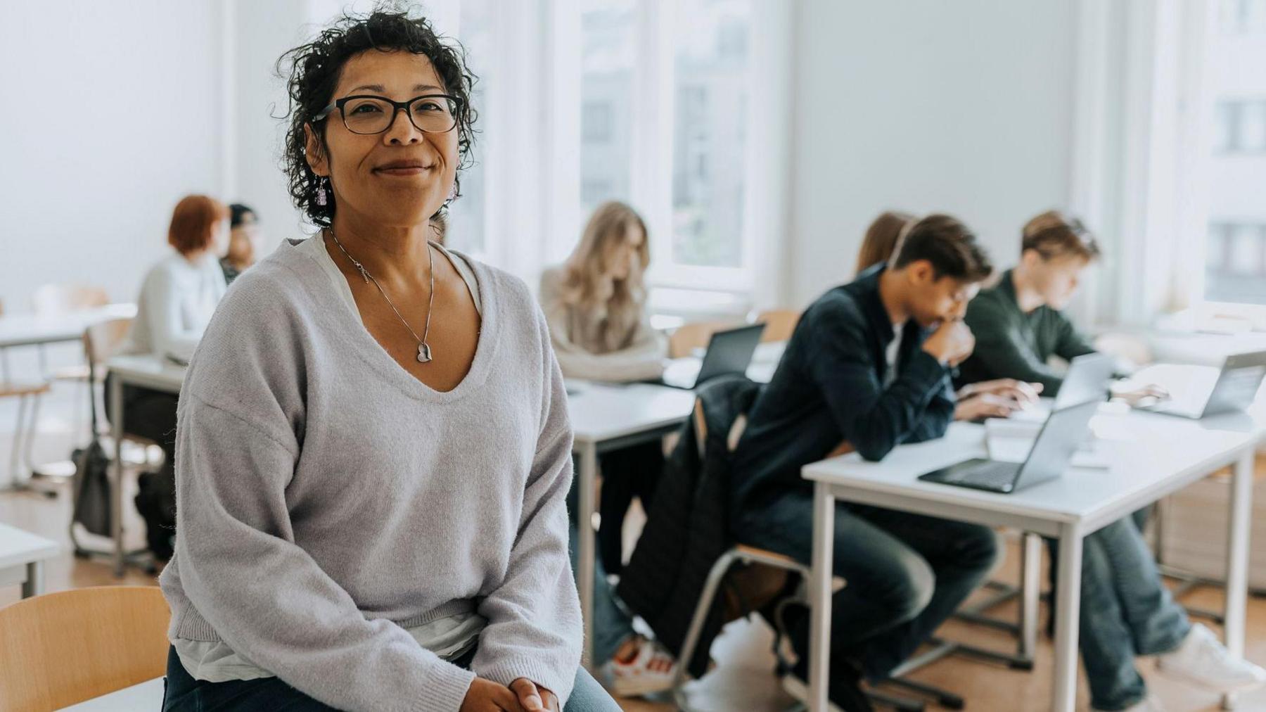 Teacher sits on a desk in a classroom with pupils working at desks behind her.