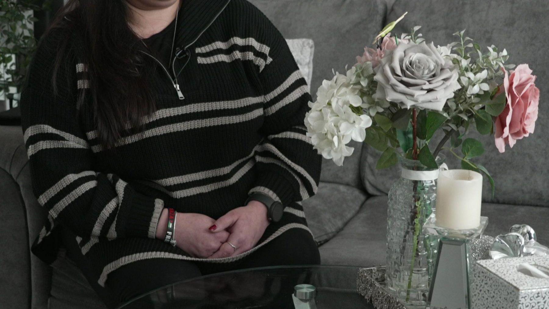 A woman with her face out of frame sits on a grey sofa with her hands clasped together in a black and white jumper. She is sat in front of a vase of flowers, a candle and a box of tissues set on a table.