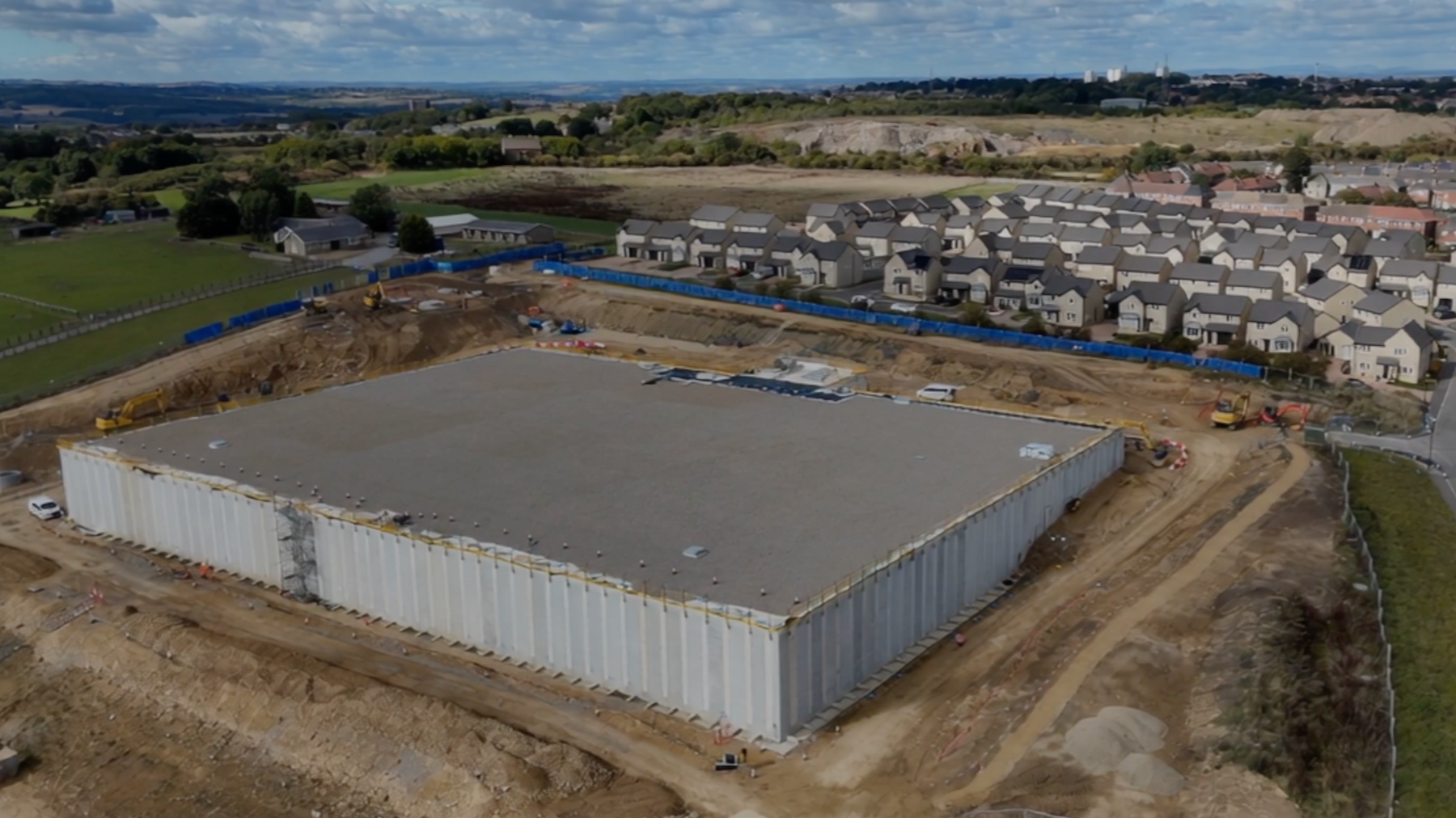 An aerial picture showing the reservoir in its final stages of construction. In the background there are several rows of new build homes, while the reservoir looks like an enormous concrete container surrounded by earthworks. It has been covered with grey gravel.