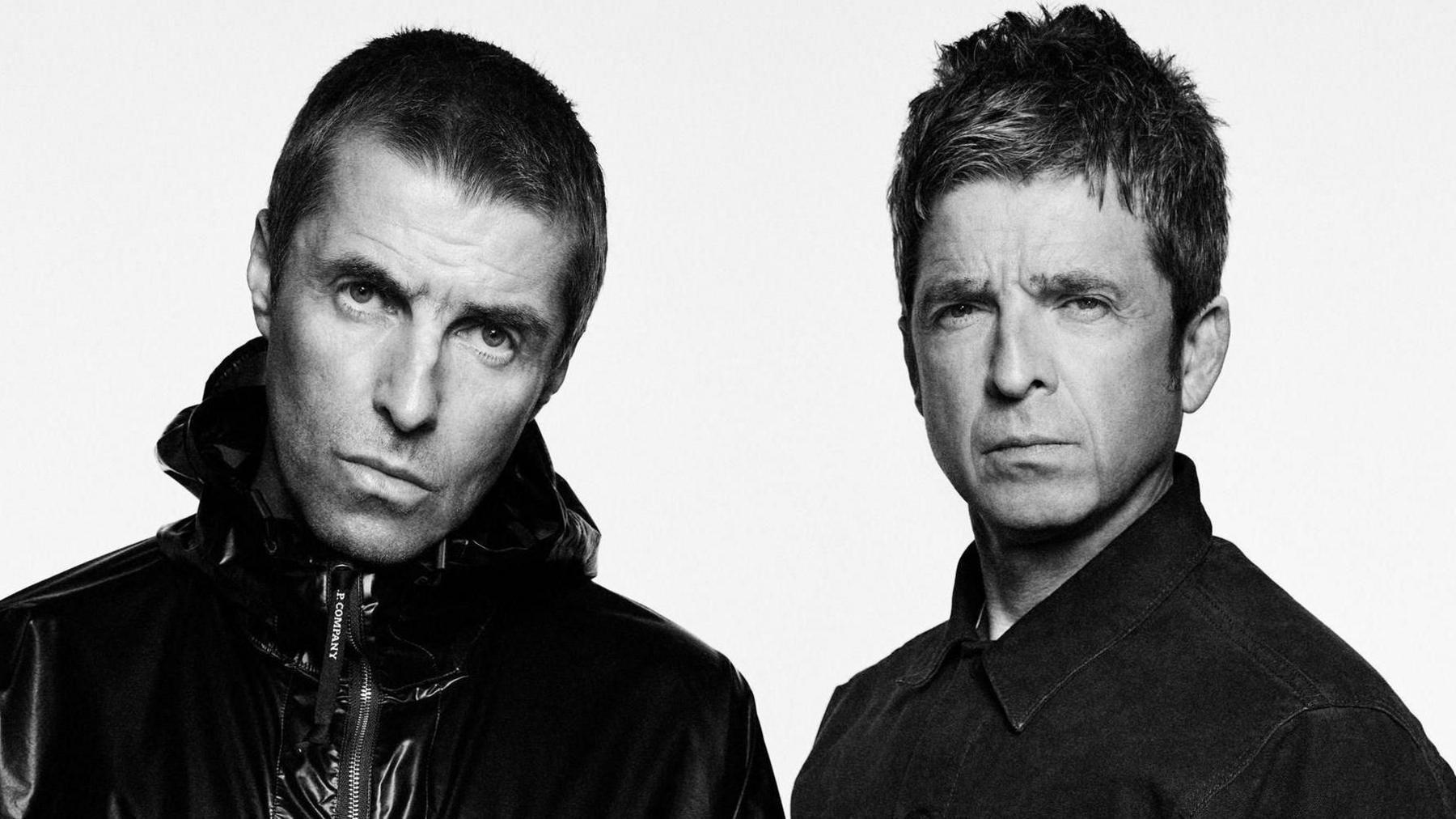 Black and white image of Liam and Noel Gallagher. Both are looking at the camera with serious faces. Liam is on the left and is wearing a shiny jacket. He has short dark hair. Noel, on the right, is wearing a denim shirt. He has short spiky hair. It is a head and shoulders shot.