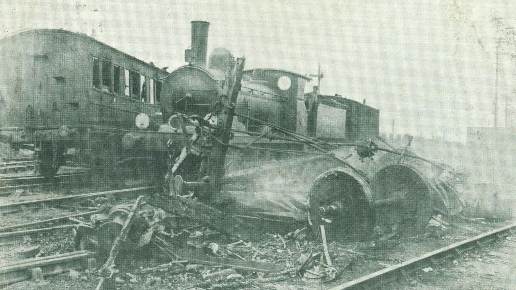 The Witham railway crash of 1905. A black-and-white image shows the remains of a railway carriage, with wheels and iron framework surrounded by broken pieces of wood, sitting on railway tracks. Smoke is rising from the wreckage, and there is a locomotive engine in the background.