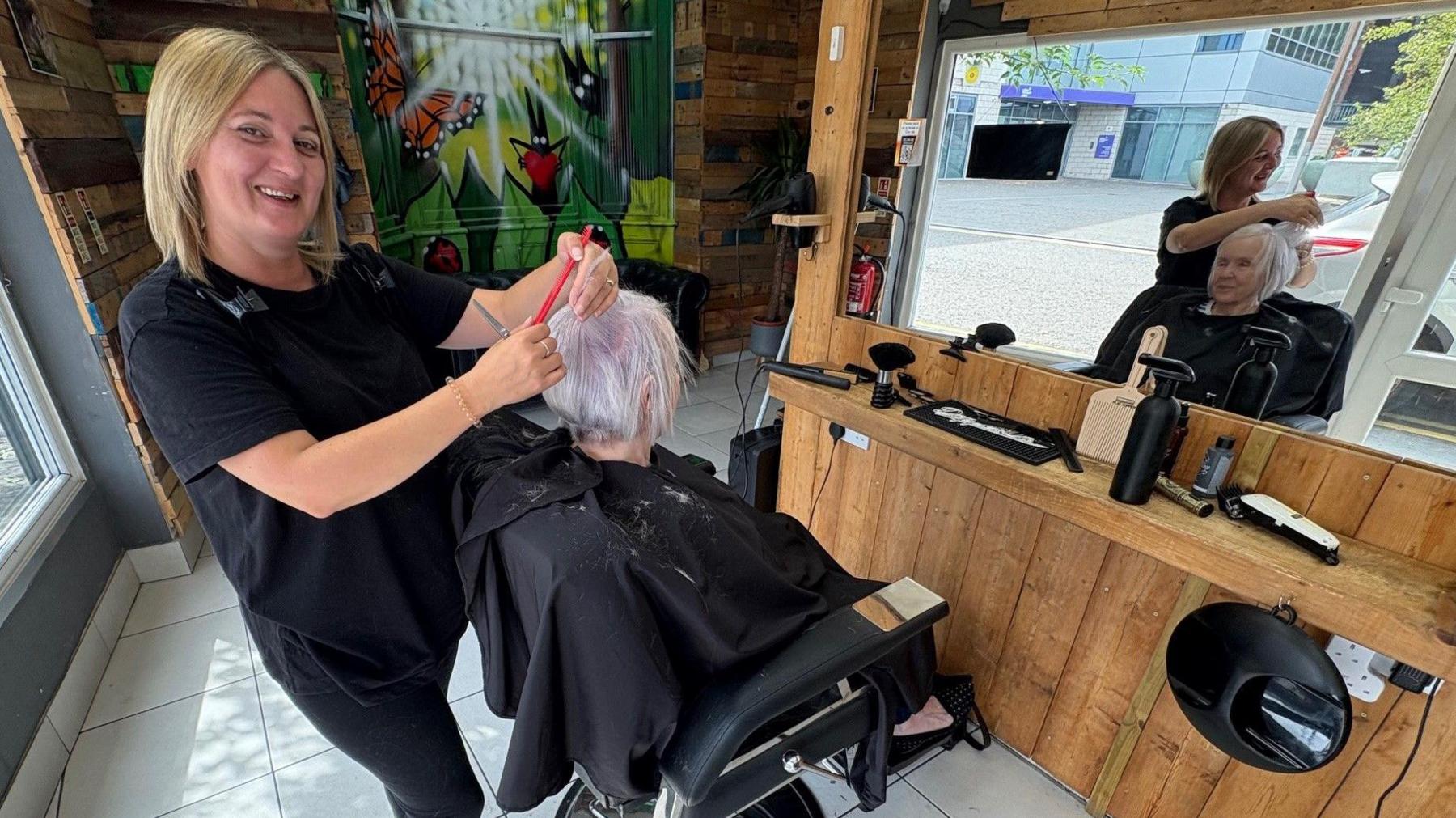 A woman with long blonde hair cuts an older woman's hair. The stylist is smiling for the camera.