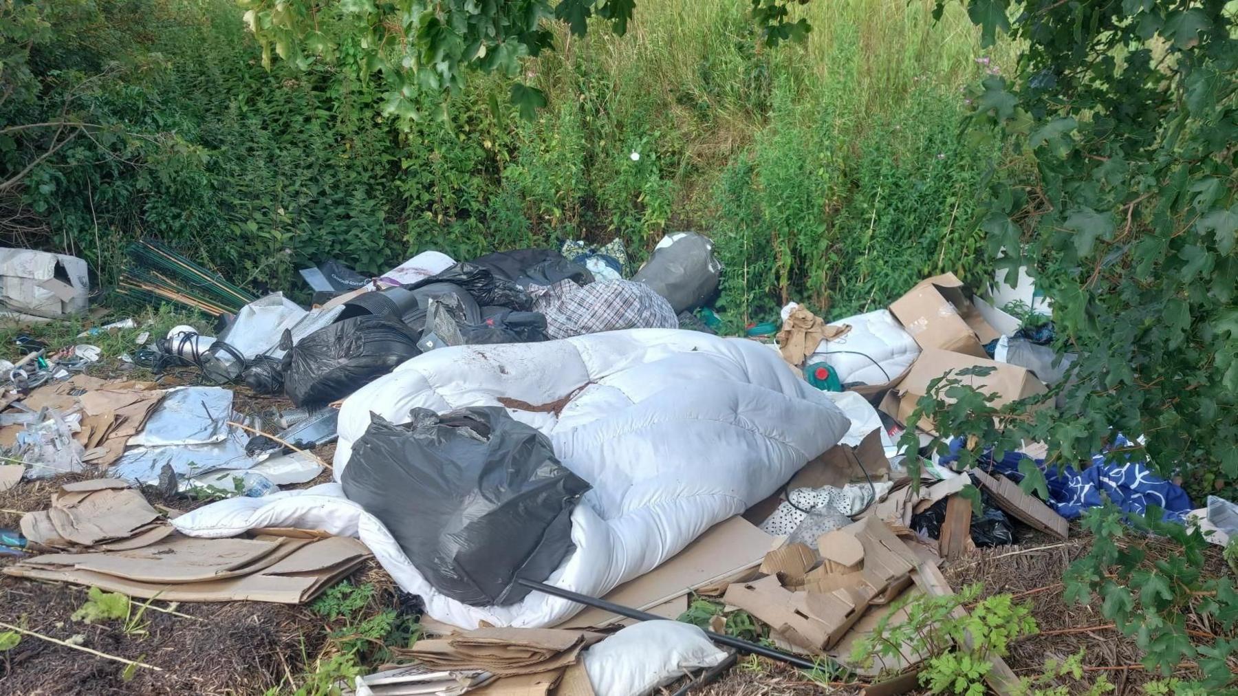 Fly-tipped items left near the bushes and nettles. It shows lots of cardboard, black rubbish bins, a white duvet and other bags and detritus.