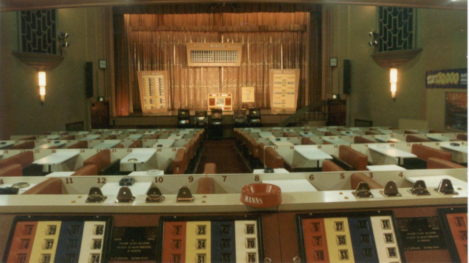 The inside of the Ritz, with a stage, all set up for bingo, with bingo counters, large art deco lights, rows of seats with tables, all numbered, with an ashtray on a counter.