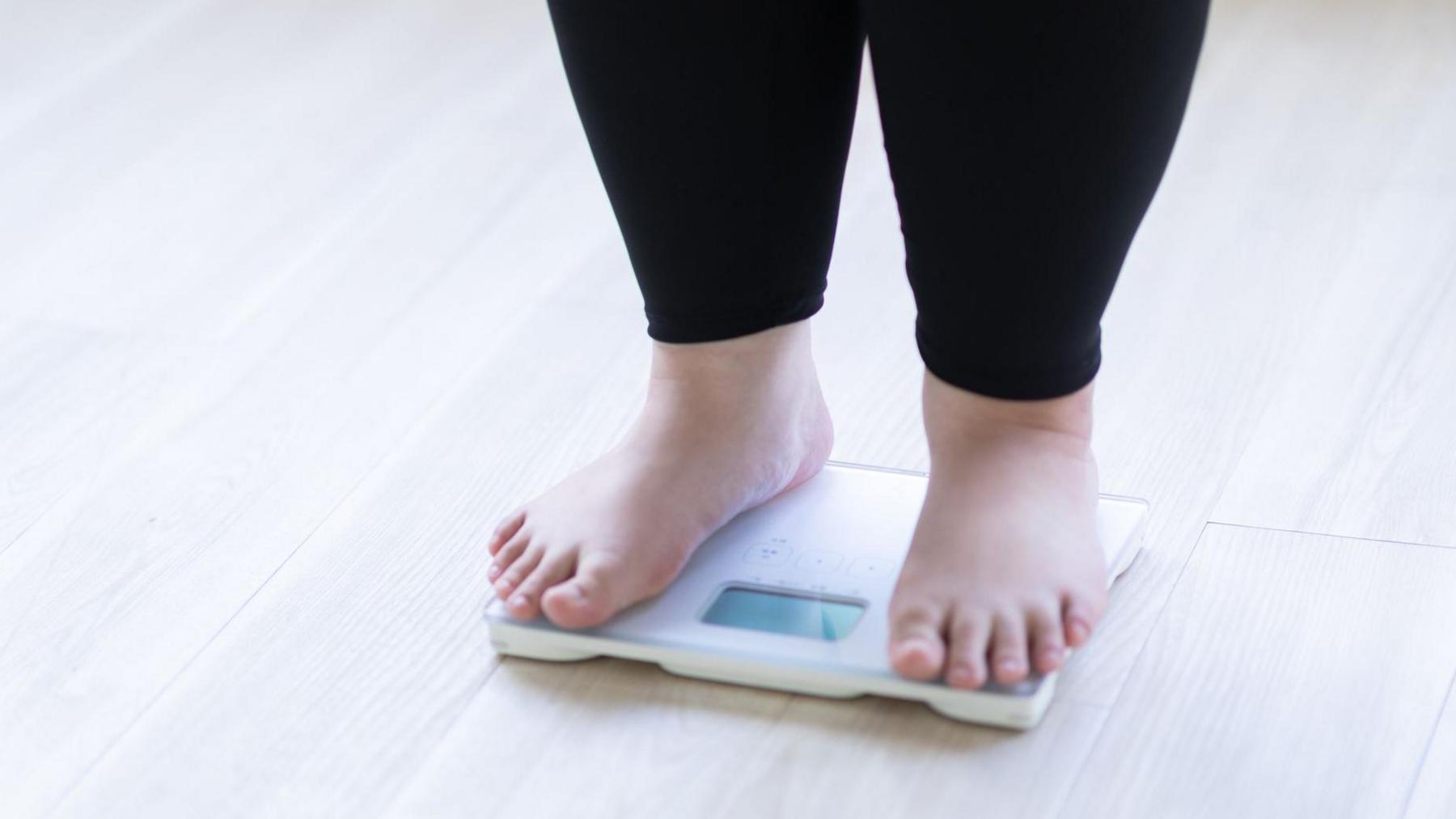 A child stands on weighing scales wearing black leggings and bare feet