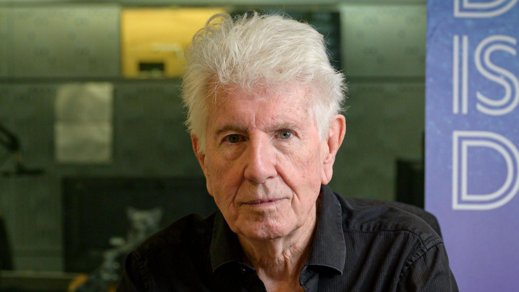 A man with white hair stares at the camera, wearing a black cord shirt, in a studio