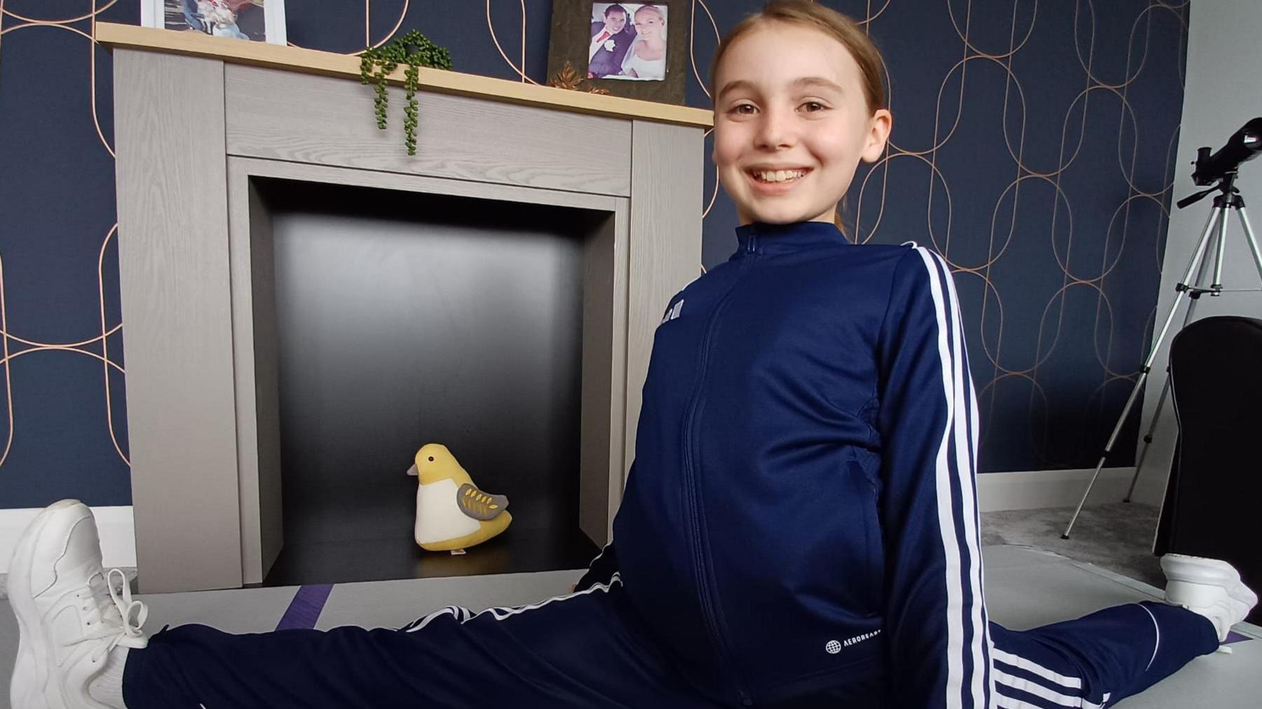 Amelia doing the splits. She's wearing a navy tracksuit and white trainers.