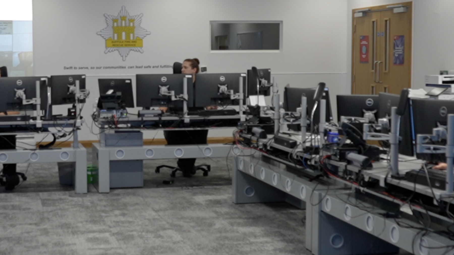 Part of a fire control room where there are rows of desks with computers and monitors on them. Some staff sit behind the desks working.