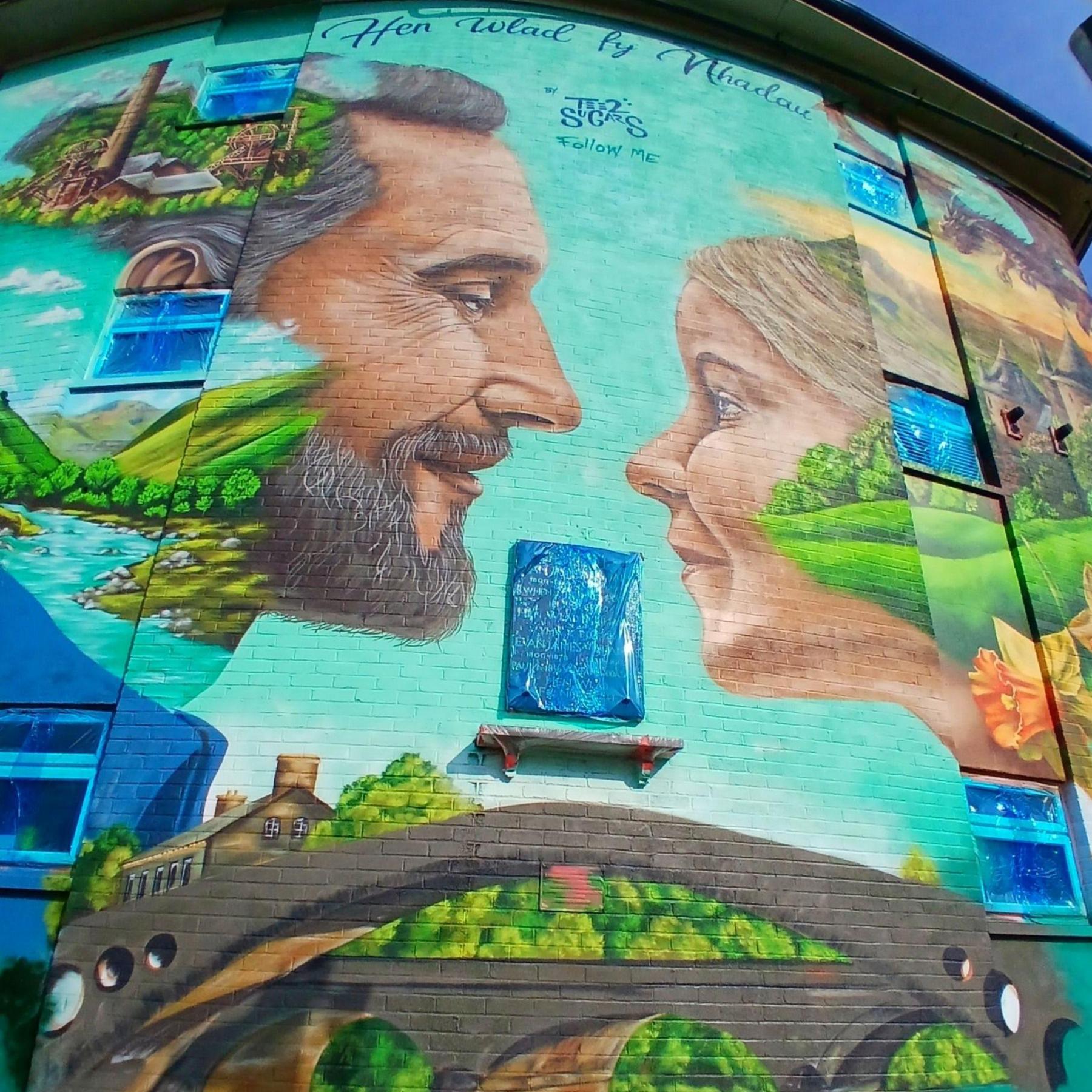 Mural promoting the eisteddfod on a wall in Pontypridd town centre showing the Old Bridge, a man and a woman smiling at each other, words from the national anthem, and scenes of mountains, valleys, and a daffodil