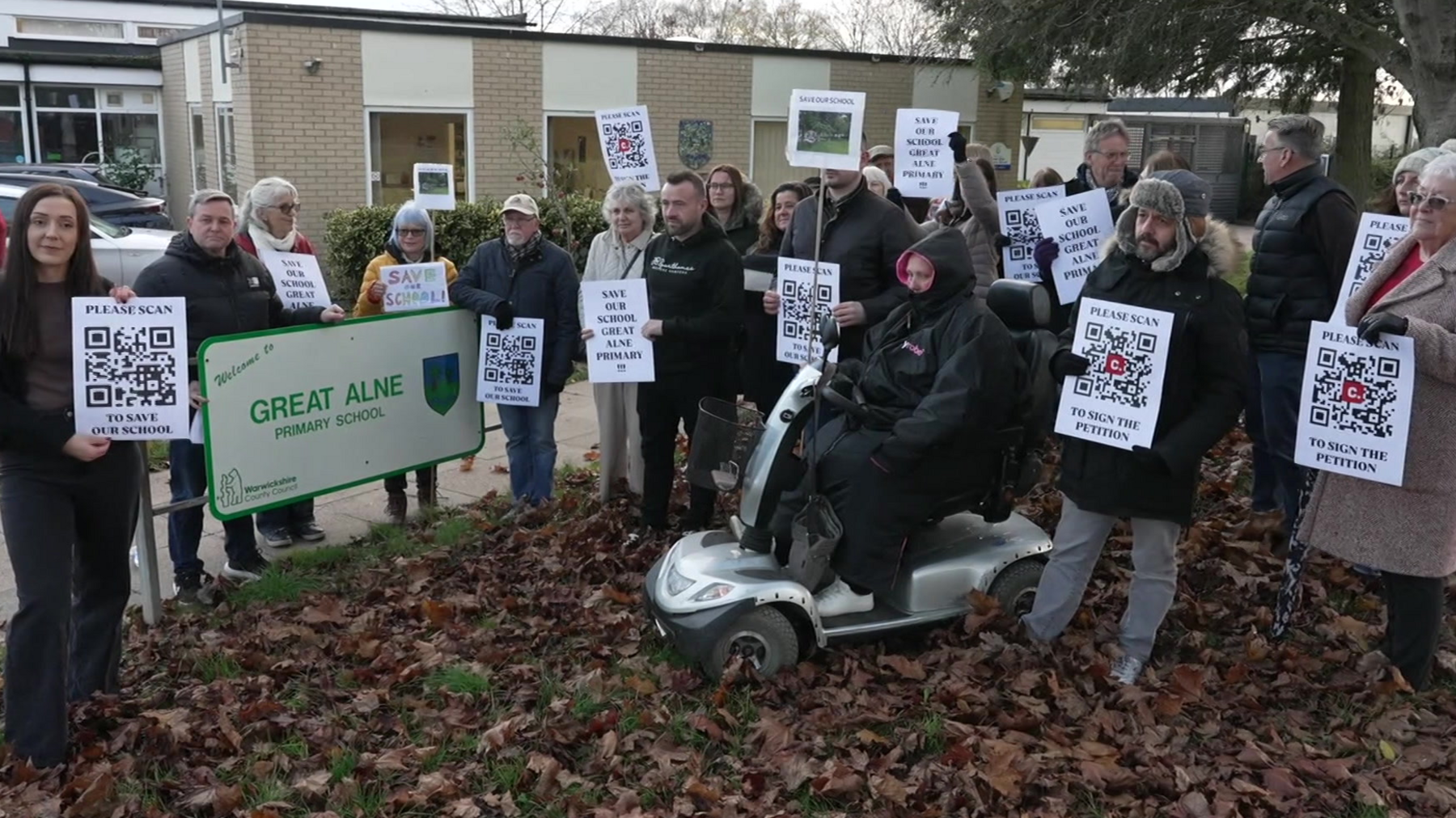 More than a dozen people are pictured holding posters and placards at the school. They're standing on leaves and grass by a sign for the school and a beige-bricked school block can be seen behind them. A person sitting on a mobility scooter is among the crowd.