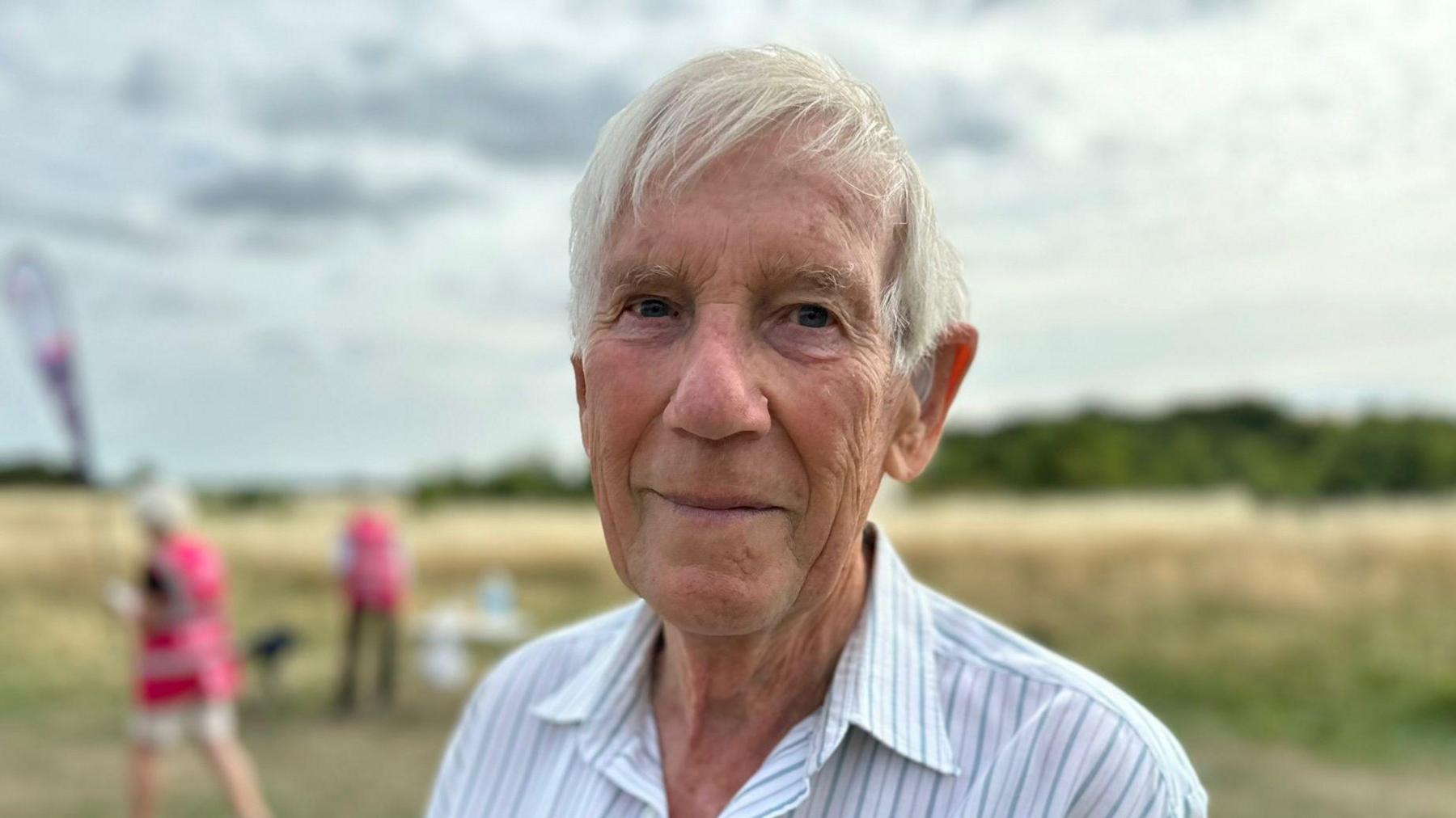 Michael Thorley has white hair and is smiling at the camera, wearing a light blue and white striped shirt. He is standing in a field, in front of some long grass. The blurred figures of people in pink tabards can be seen behind him.