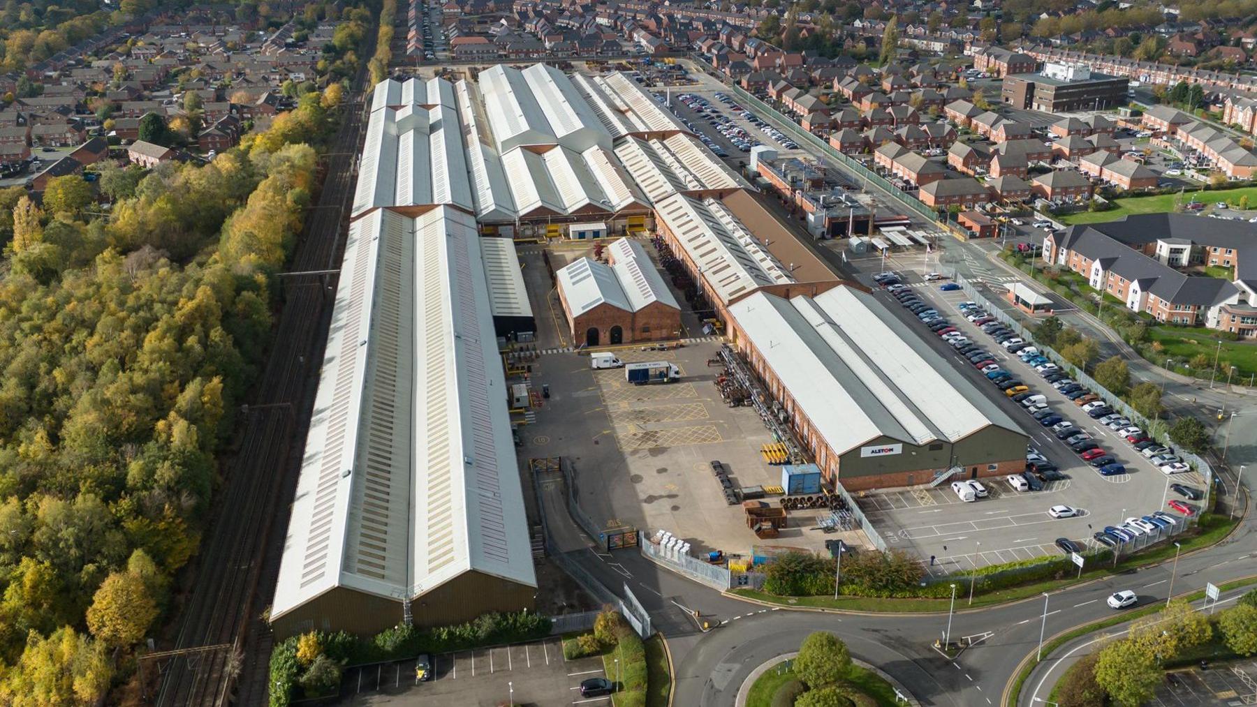 An aerial view of the Crewe Works factory - a series of buildings and car park. There is a railway track on the left hand side of the image and a car park on the right hand side. In the middle of the closest set of buildings is a goods yard.