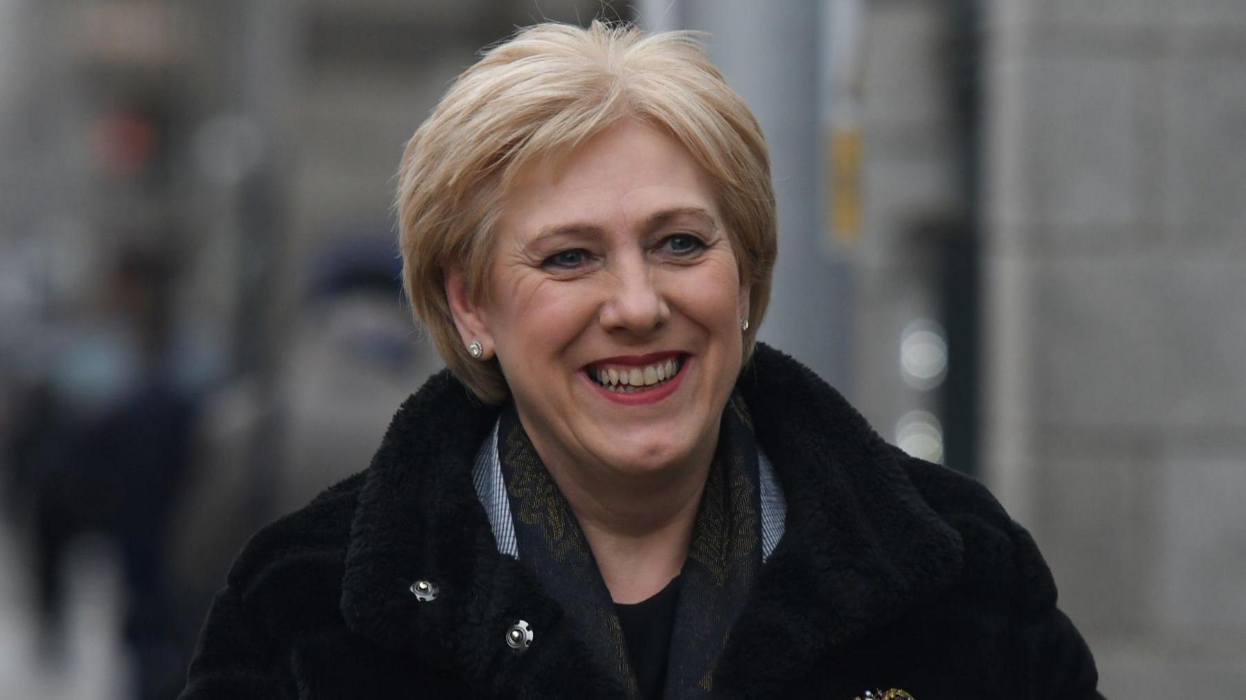 Heather Humphreys walks outside Leinster House. She is an older woman with short, blonde hair and she is smiling as she walks. She is wearing a puffed black coat and a thin scarf.