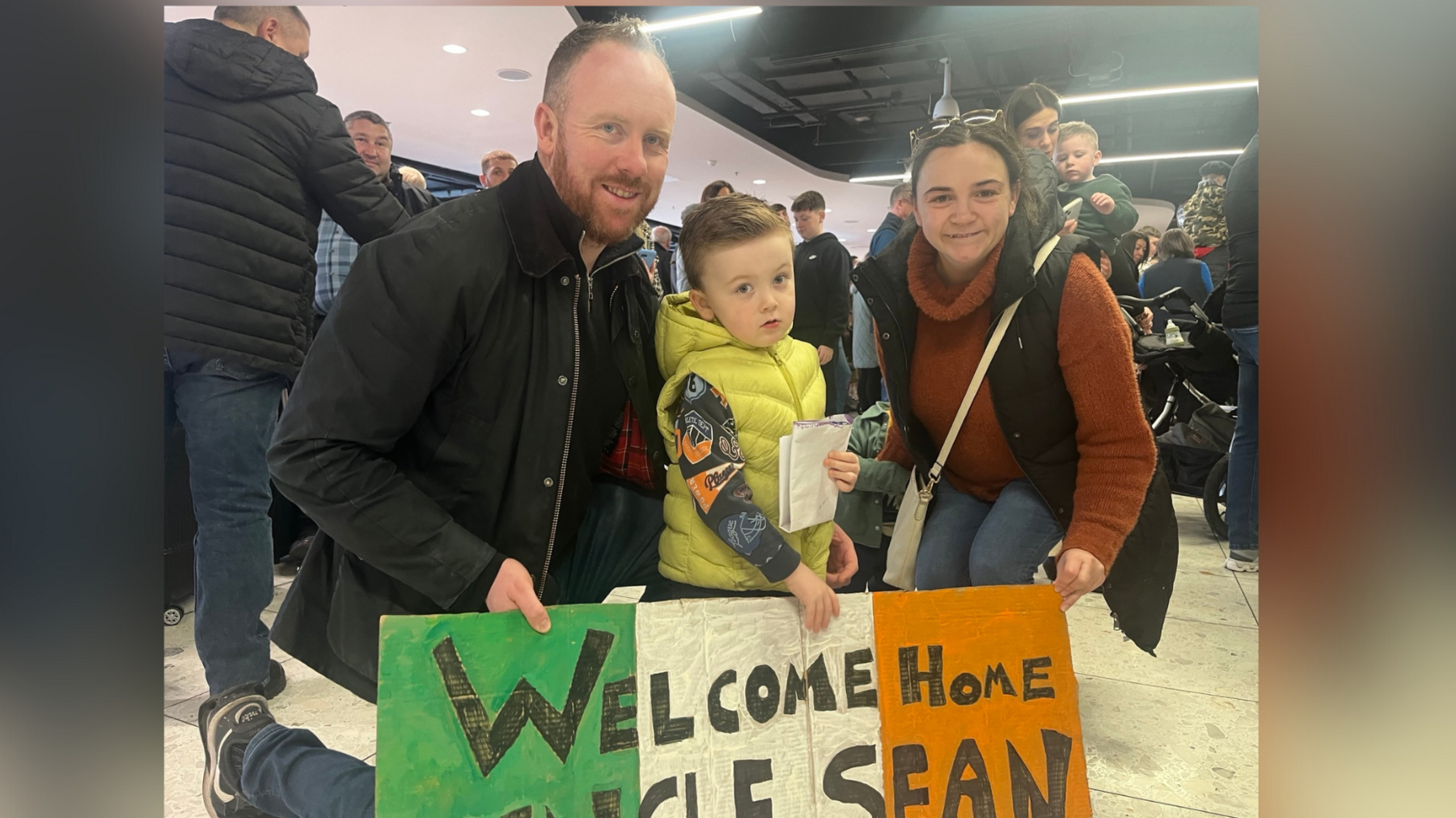 A man dressed in black, a young boy in yellow and a woman in an orange top with a black body warmer smile as they hold up a green white and orange sign which says - Welcome home uncle Sean.