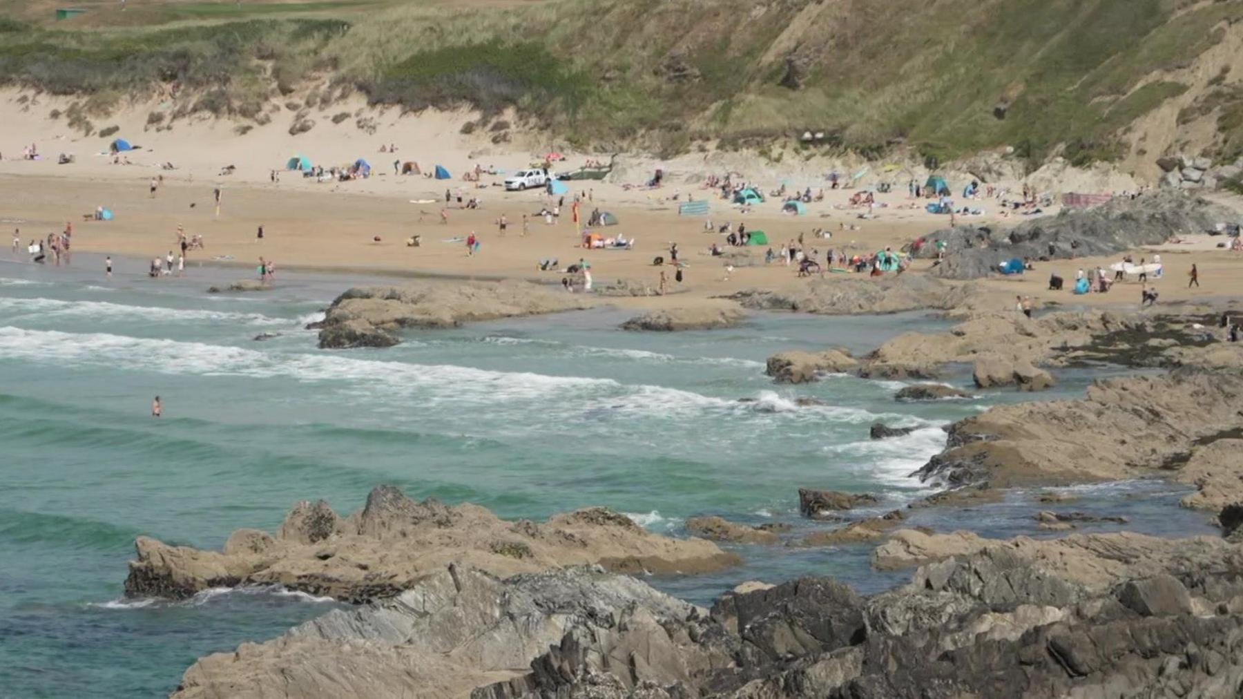 Fistral Beach is a wide sandy beach with lots of people walking and lying on it and there are people swimming in the sea alongside jagged rocks. There are many windbreaks and beach tents. It is a slightly cloudy day.
