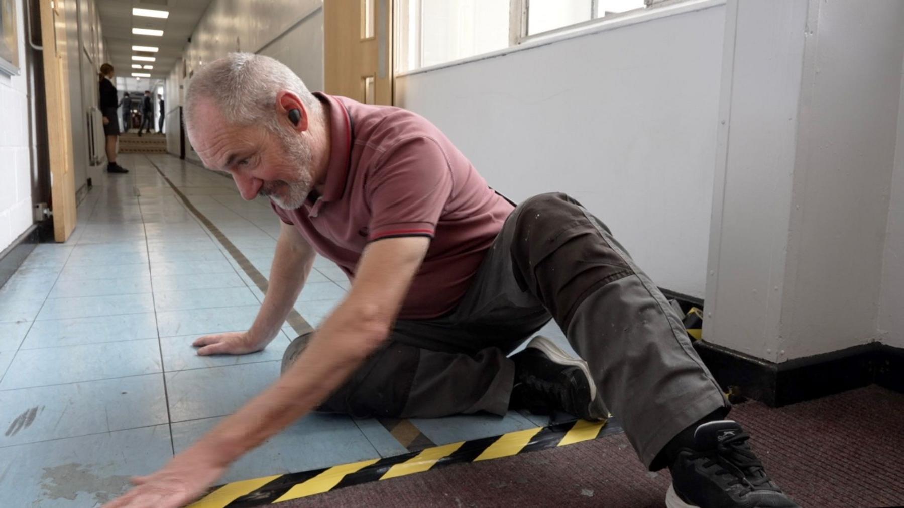 A man wearing a red polo shirt sticks yellow and black tape along the edge of a piece of carpet in a corridor at Patchway Community School.