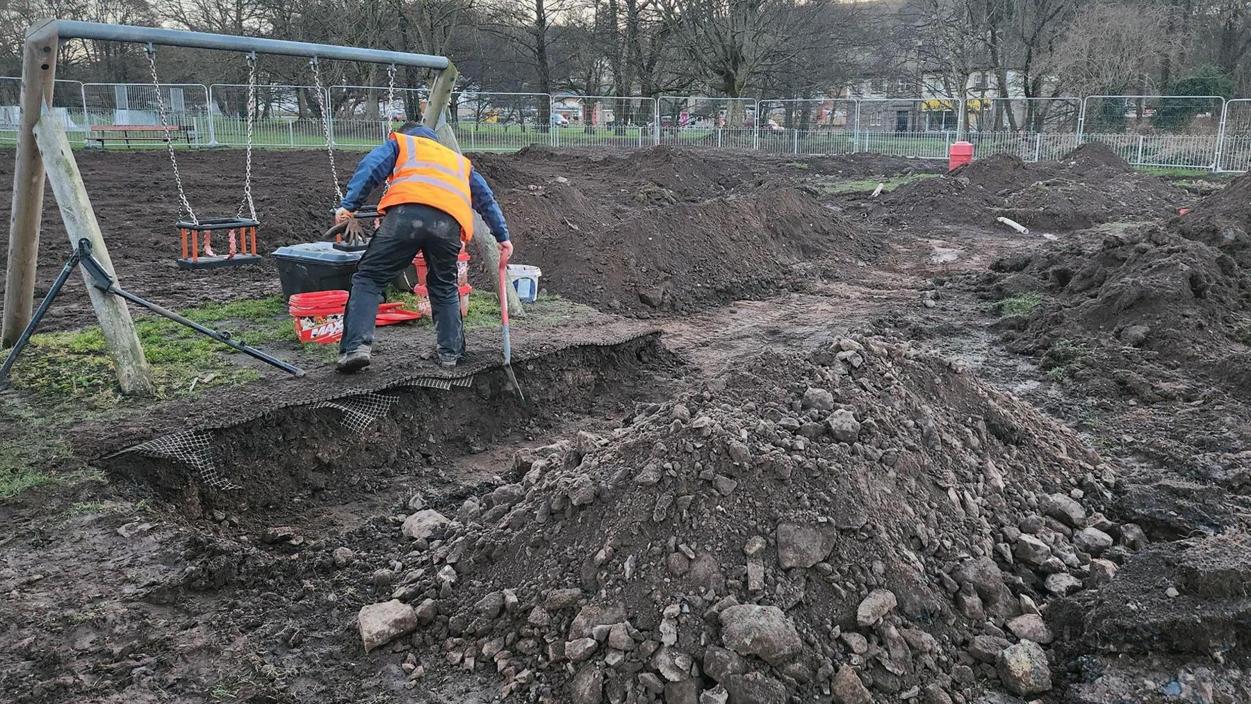 A children's park has been completely dug up with numerous piles of earth and metal fencing all around the site. Just one swing remains standing, a man is digging in front of it. 