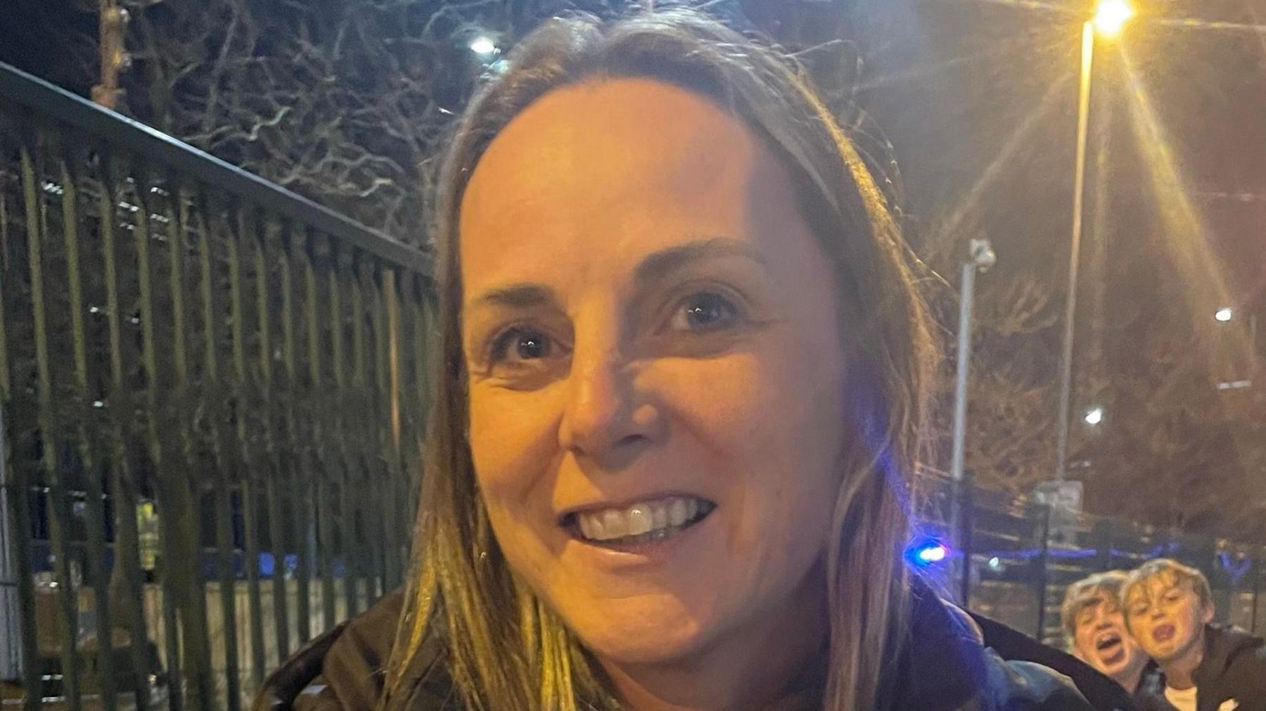 Plymouth Argyle fan Kate smiles as she leaves the Etihad Stadium where she watched her side lose 3-1 to Manchester City in the FA Cup fifth round. It is night time and two boys are standing sticking their tongues out behind her.