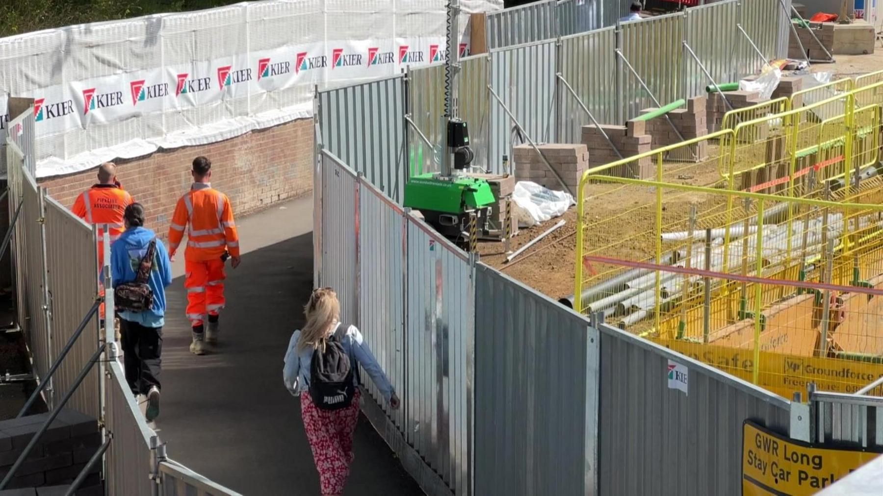 Pedestrians, including two workers in high vis clothing, walk along a pedestrian walkway in the Oxford Station works