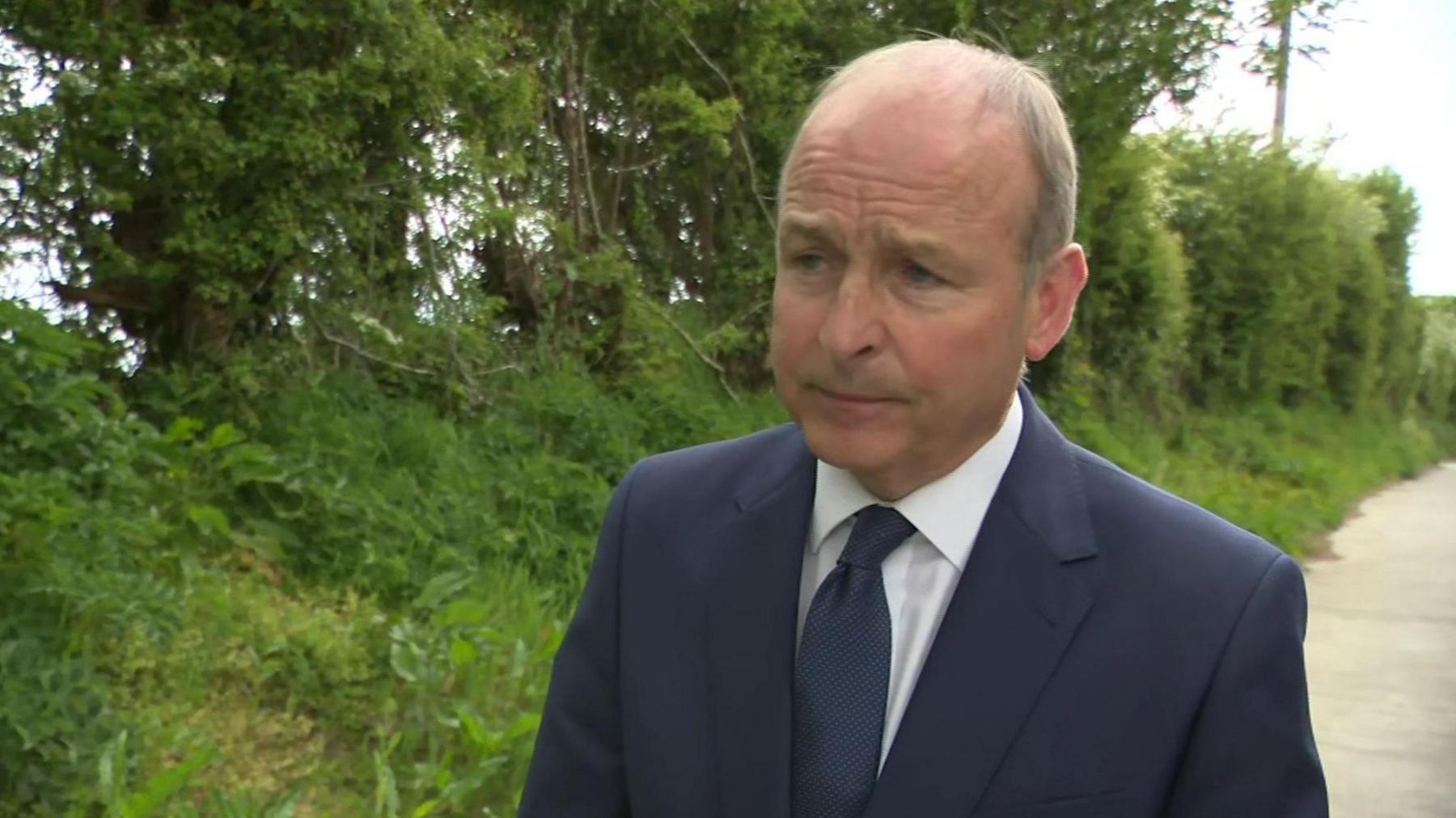 Taoiseach (Irish prime minister) Micheál Martin wearing a blue jacket, blue tie and white shirt, is seen standing at the side of a country road with trees and grass on the layby as he speaks to RTÉ News following the death of Garda (Irish police officer) Kevin Flatley