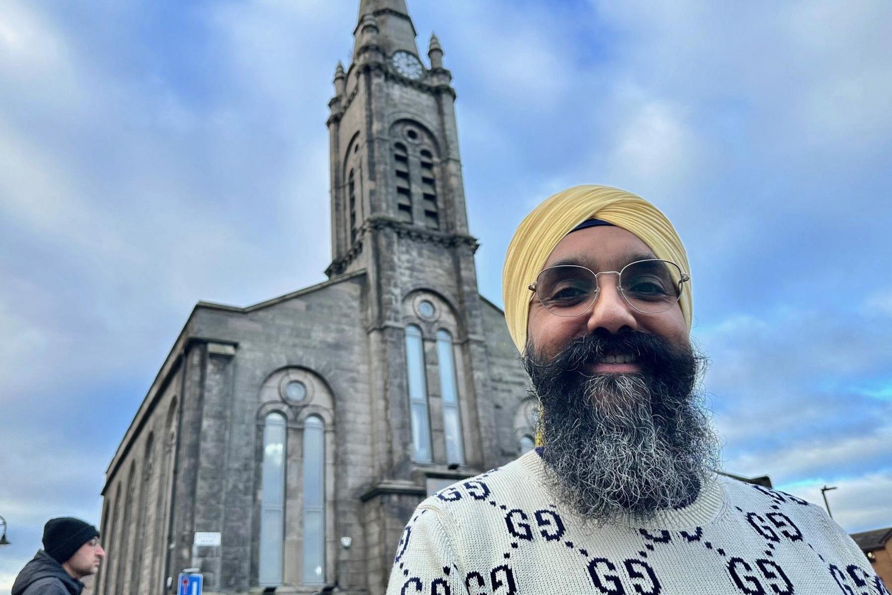 Galab is wearing a yellow turban. He has a beard and glasses. In the background is the stone built gurdwara.