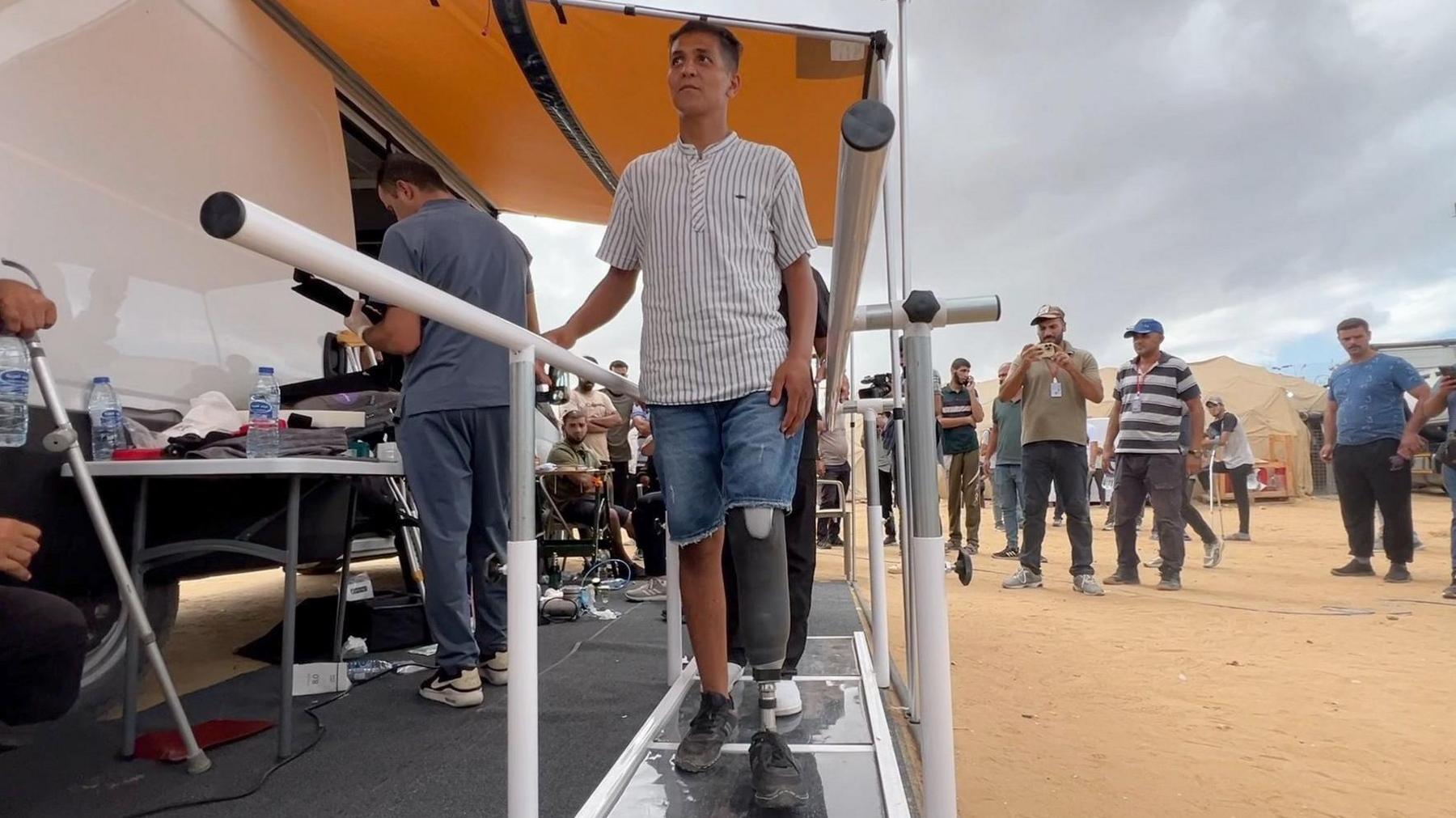 Rizeq Tafish walks using his new prosthetic leg along a special platform with long handles with dozens of people milling around him