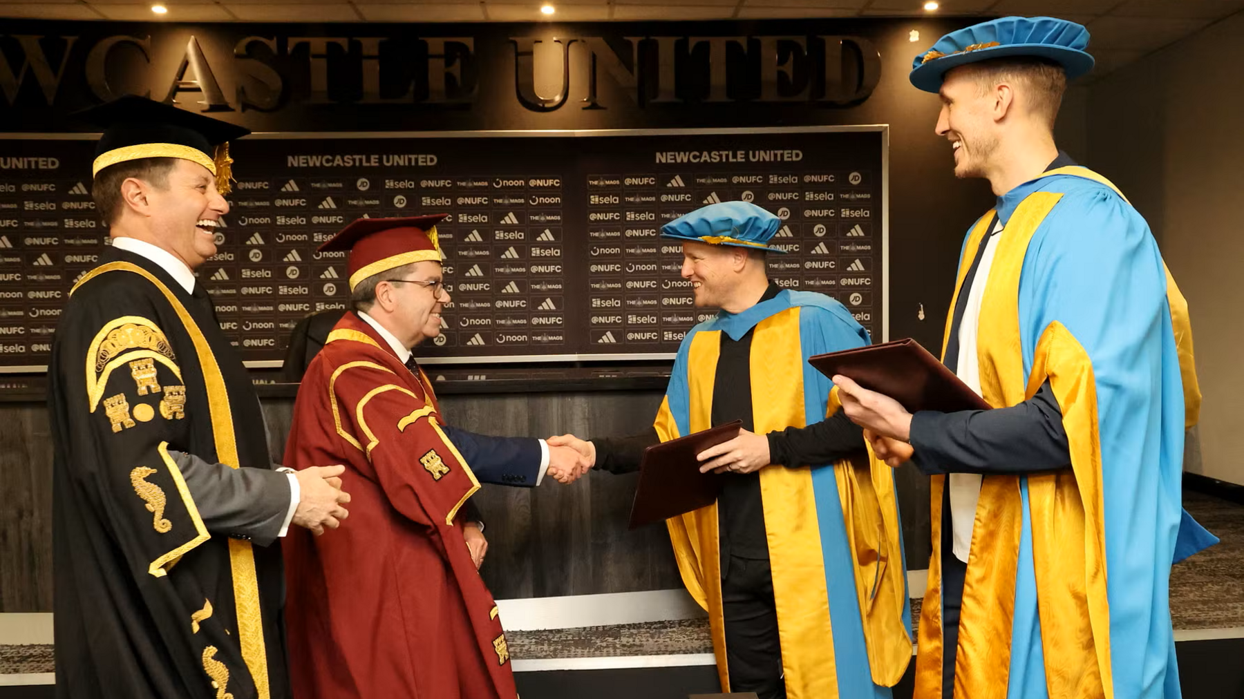 Eddie Howe and Dan Burn with staff from Northumbria University in the Newcastle United media room. The pair are wearing blue and gold robes. University chancellor George Clark is in a black robe and another man is in red, to his left. Howe is shaking hands with the man in red and smiling. They are standing in front of a black and white advertising board with various Newcastle sponsors and a long black desk.