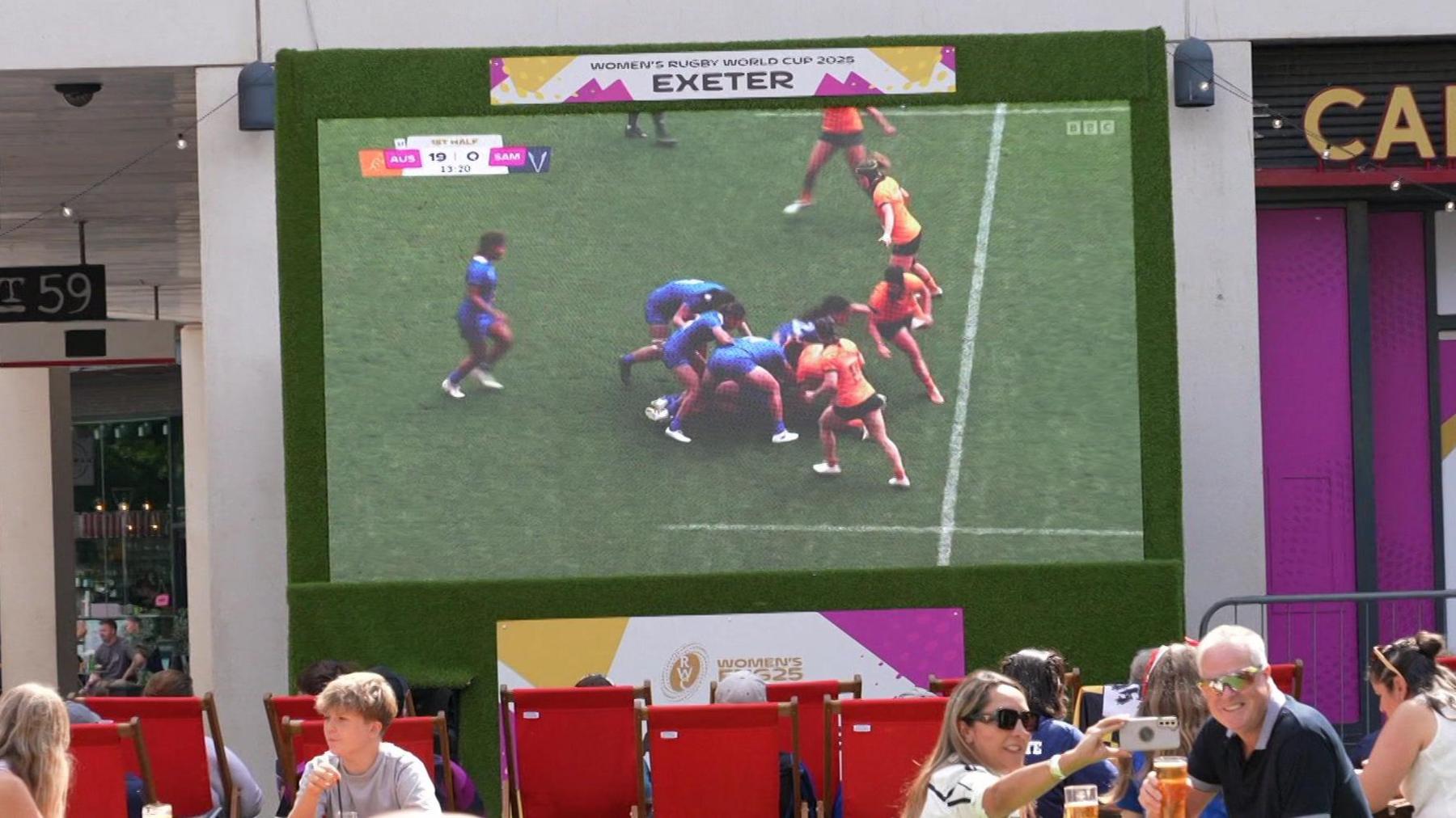 People sit on benches and red deck chairs in the official fan zone in Exeter city centre to watch Australia play Samoa at the 2025 Women's Rugby World Cup. A couple are taking a selfie while holding pints up. The score in the match is 19-0 to Australia.