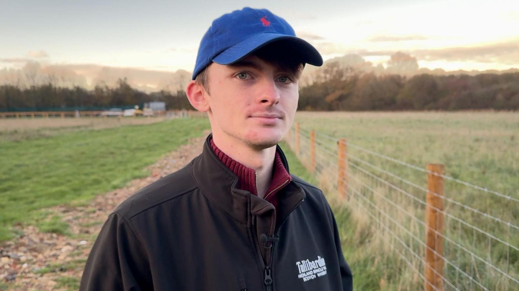 Joshua wears a blue cap and a black fleece. He stands by the fence at the edge of his field.