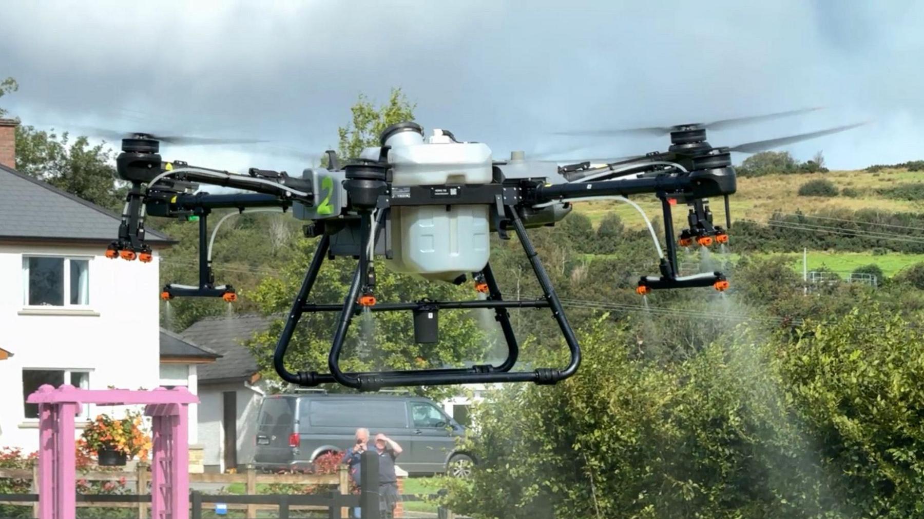 A spraying drone lifting off from a garden and spraying water. The drone is black on the four arms and under carriage. In the middle is a silver box.