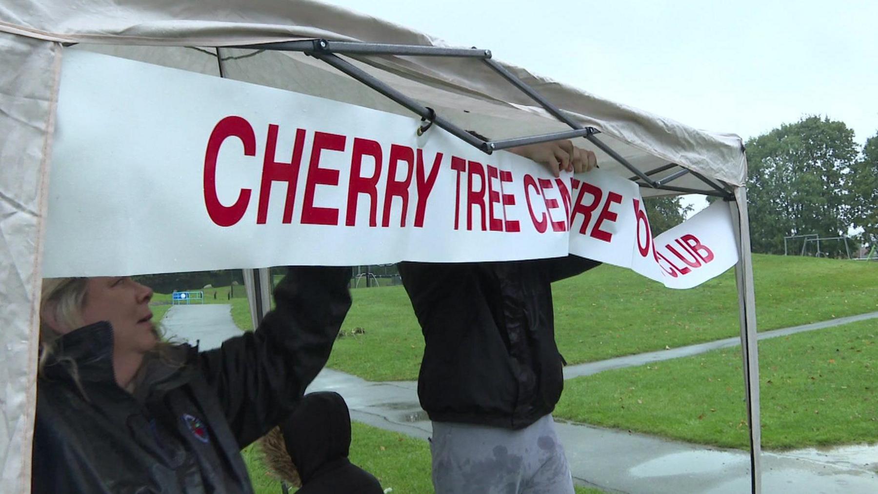 A woman and a man wearing rain coats attach a white banner, with red letters reading "Cherry Tree Centre Youth Club". to a small gazebo in a park in the rain. 