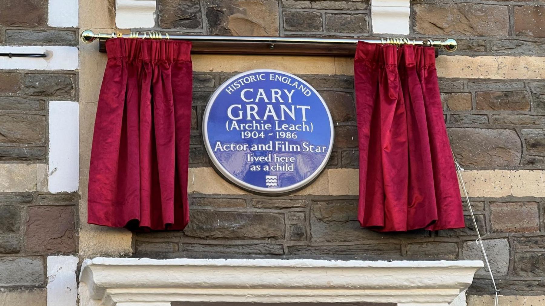 A blue plaque created in memory of Bristol actor Cary Grant is seen between two thick red drapes, at its unveiling. The plaque reads "Historic England. Cary Grant (Archie Leach) 1904-1986 Actor and Film Star lived here as a child." The text is in white writing on the blue background of the plaque