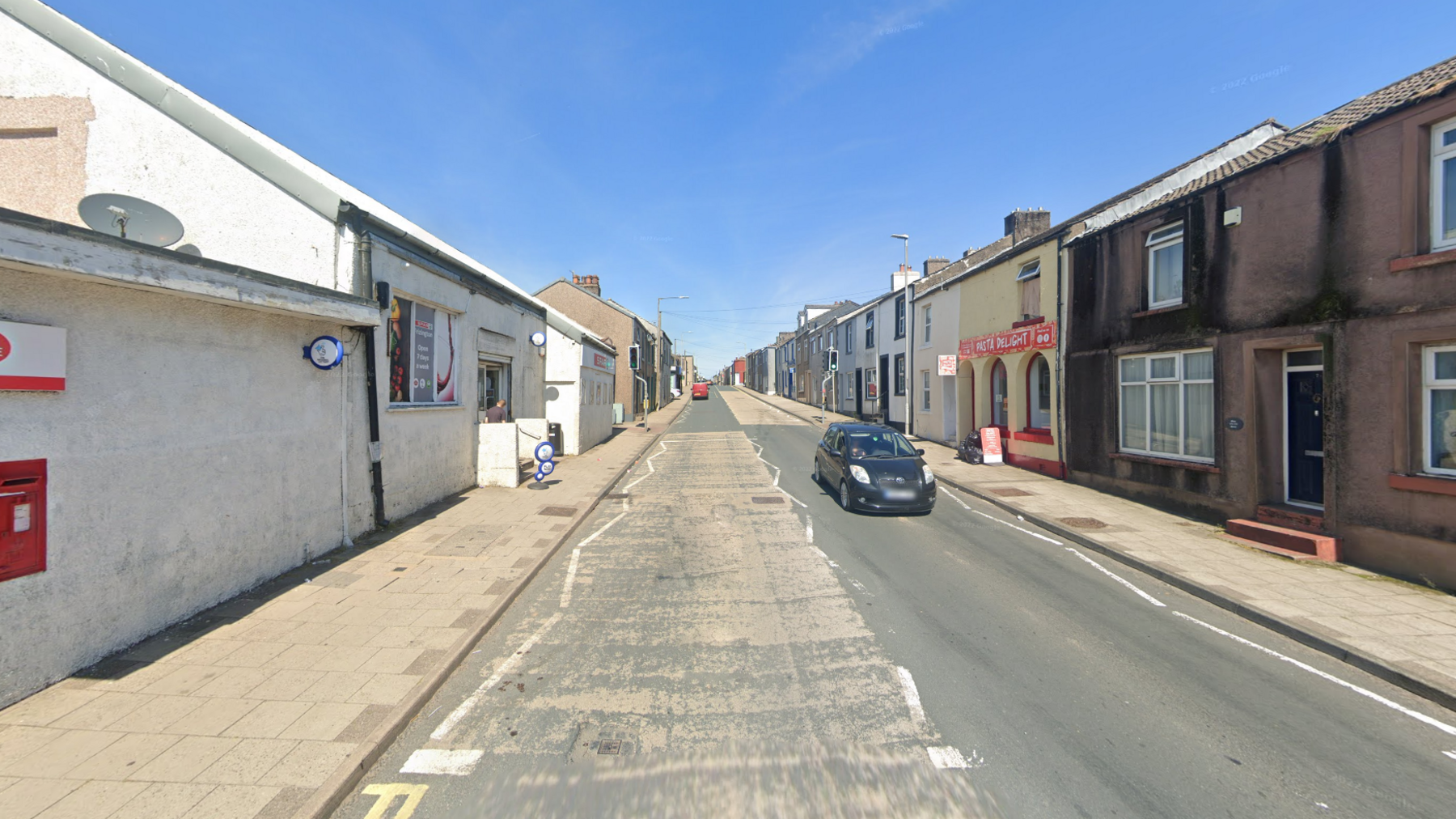 The A5086 is a single carriageway bordered by two-storey terraced houses. A post office and corner shop sign is on the left, there is a store front called: "Pasta Delight" on the left.