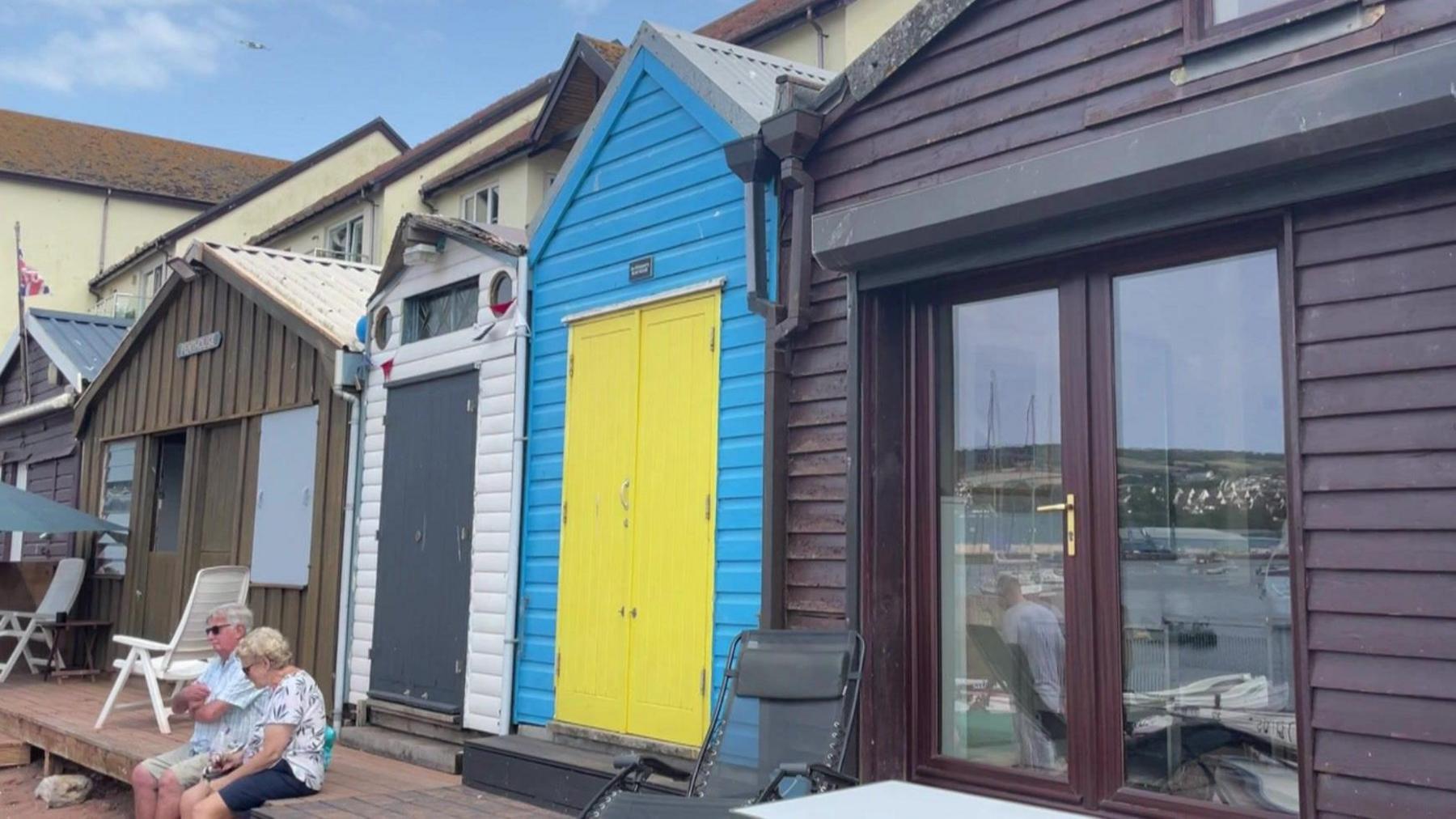 A row of beach huts including one with a yellow door and blue walls. A man and woman are sat in front of it