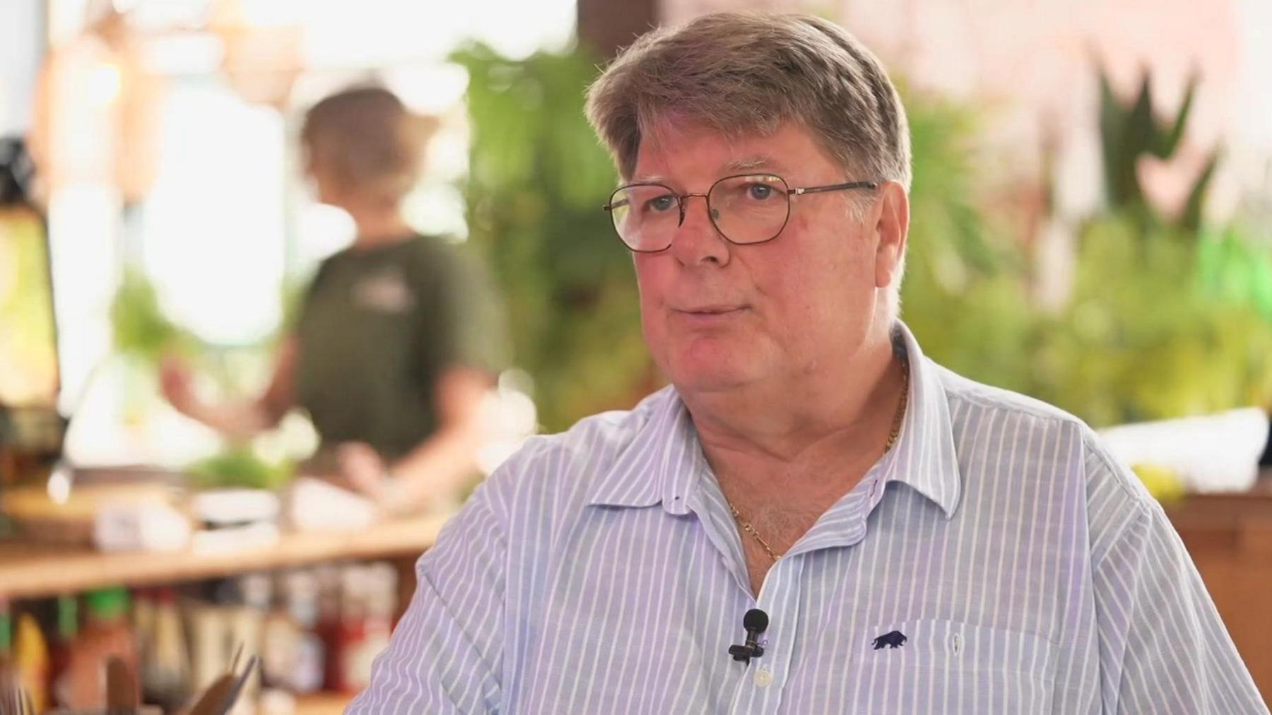 Rob Stander, wearing a blue and white striped shirt and glasses. The background is blurred behind him. There is a woman sitting at a table in the background.