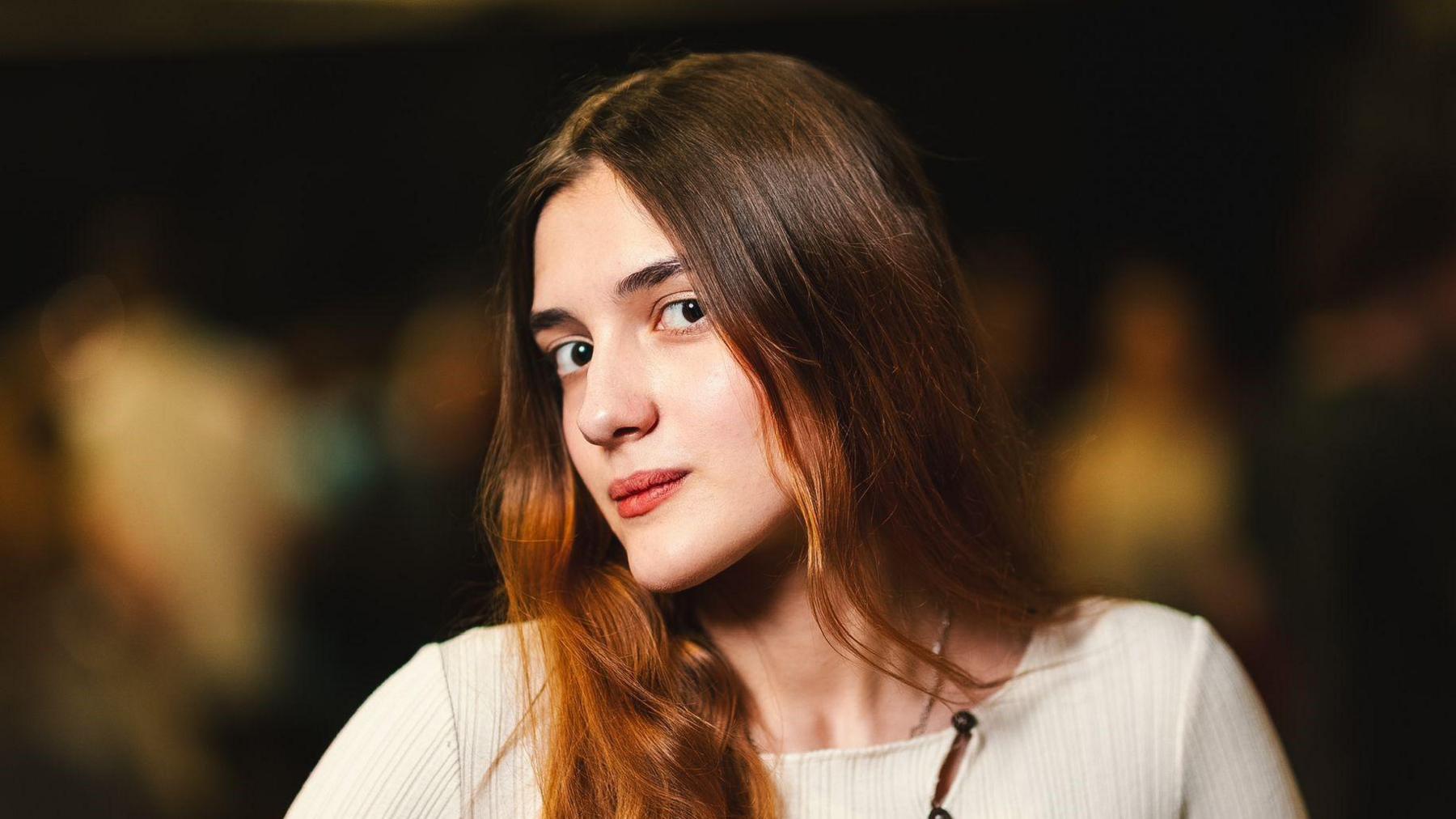 Young woman with long brown hair and beige top with turned head looking into the camera. Blurred background.