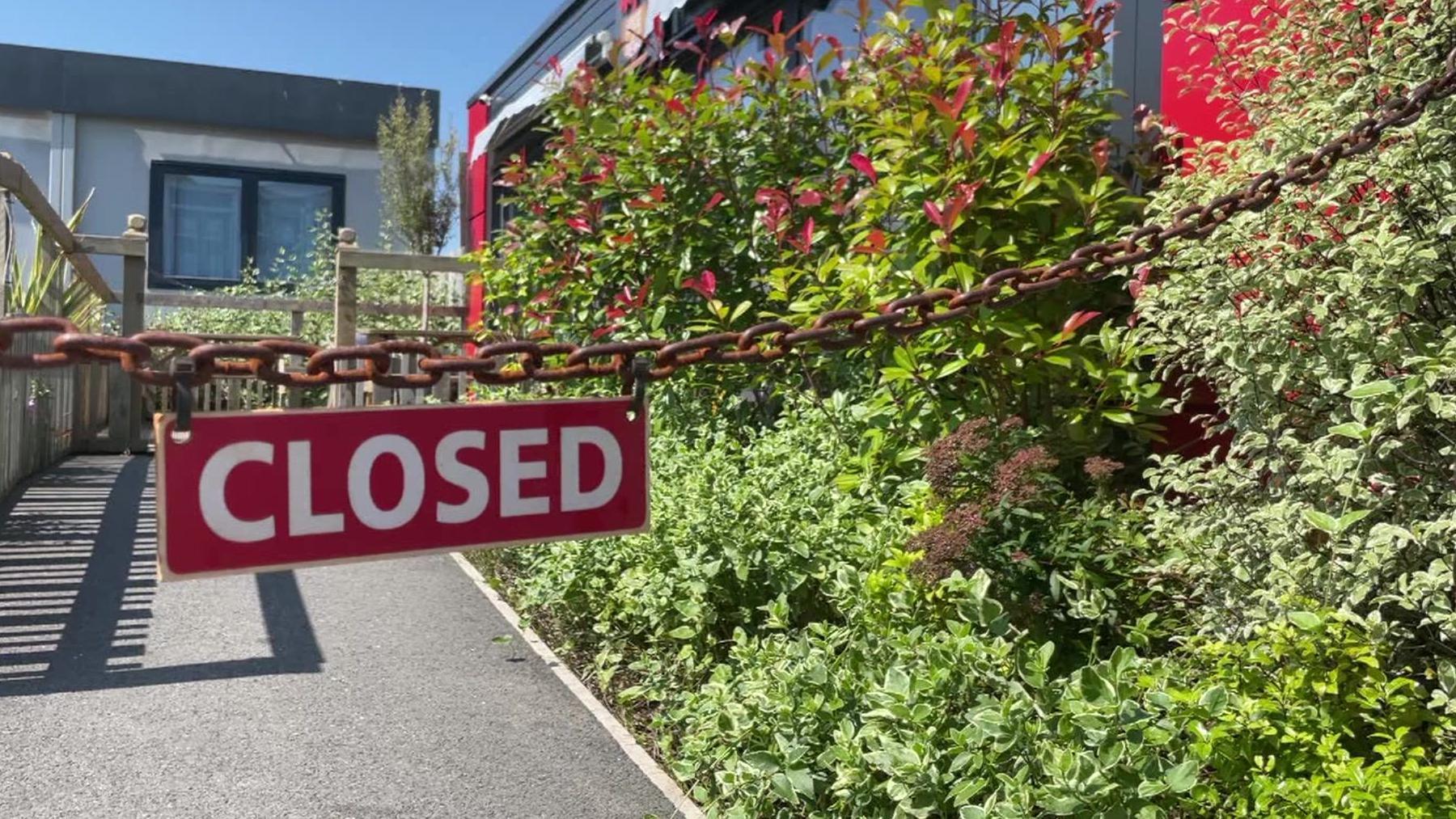 A closed sign with white letters and a red background hanging from a chain in front of a shop unit and a set of plants on a sunny, cloudless day.