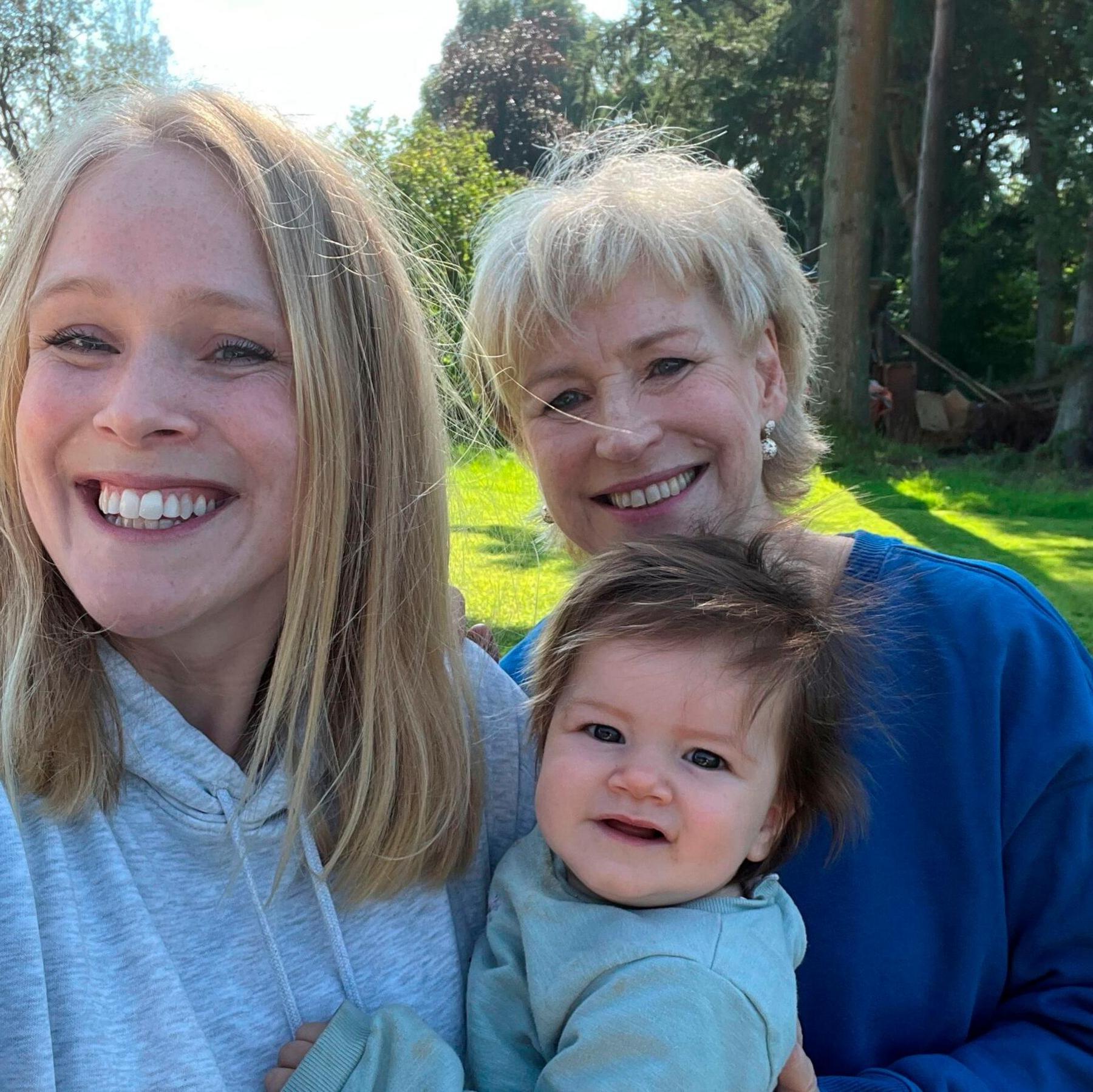 Selfie of Sally Magnusson (right) in a navy blue top next to her daughter Anna Lisa who is wearing a grey hoodie. Anna Lisa is holding her daughter Remy who has dark hair. The sun is shining on the bright green grass and trees behind them and they are all smiling at the camera.