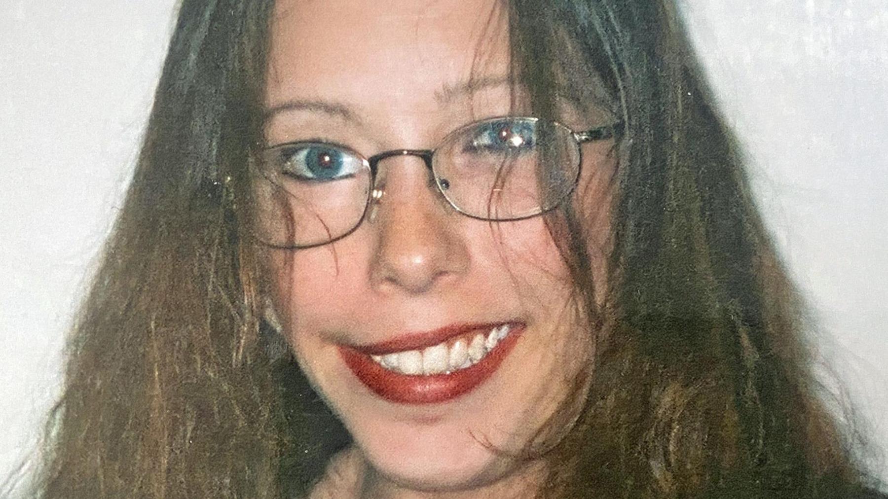 A woman with long brown hair, blue eyes and glasses smiling at the camera