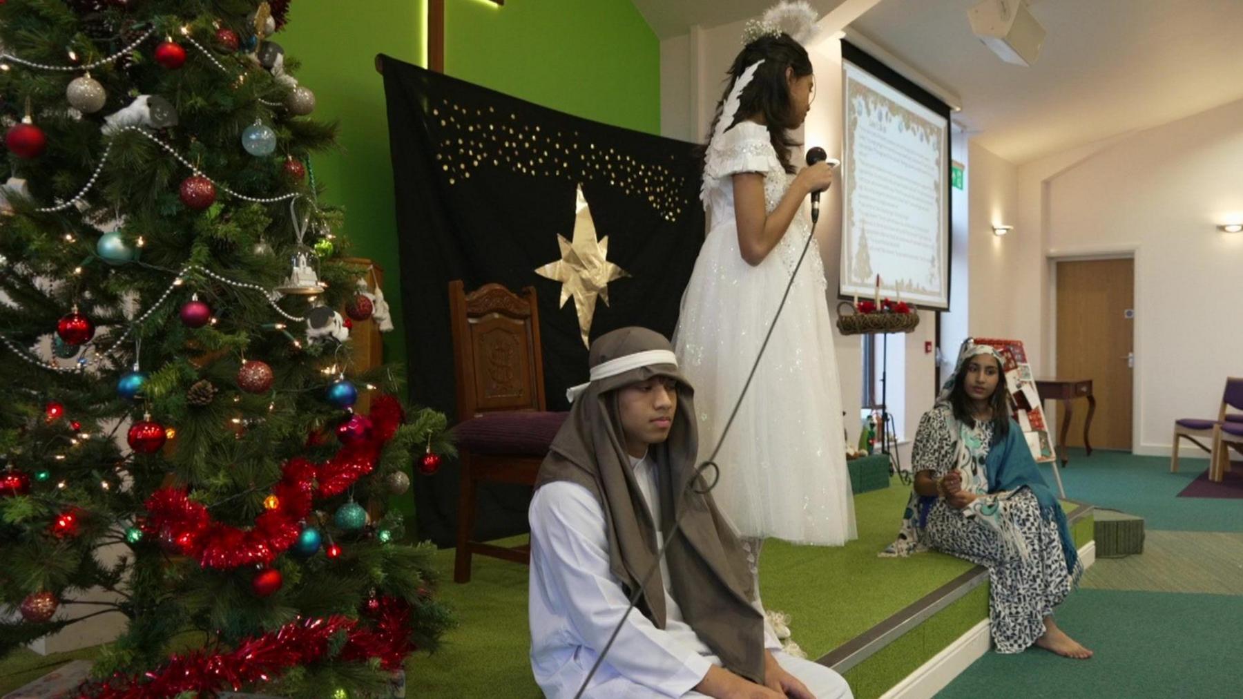 A girl dressed as a angel in a white dress and wings stands on a small stage between a boy dressed as a shepherd and a girl in a blue headscarf.