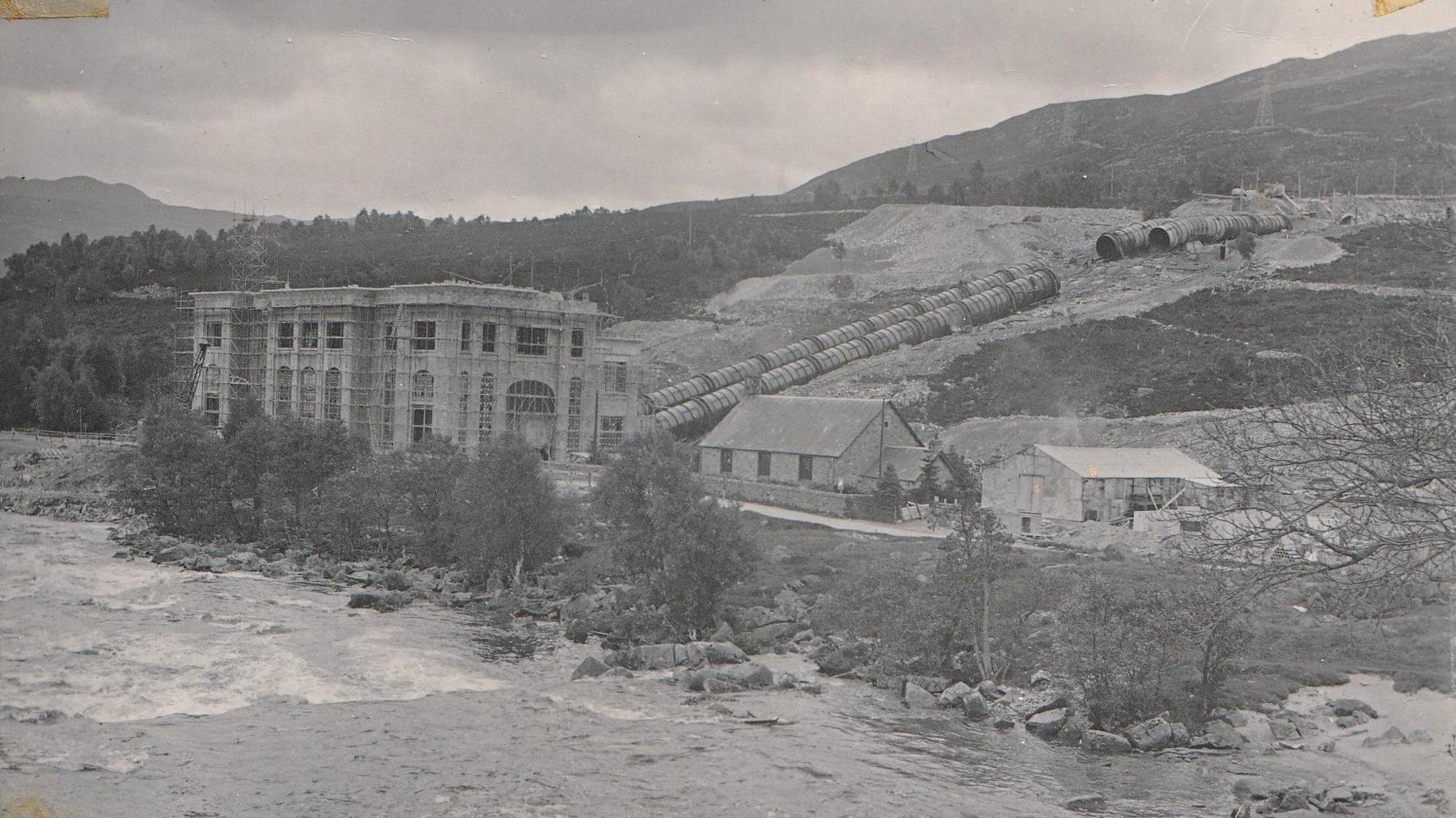 The Tummel power station while it was under construction. It is covered in scaffolding, with large pipes coming down from the hill behind and a fast-flowing river in front