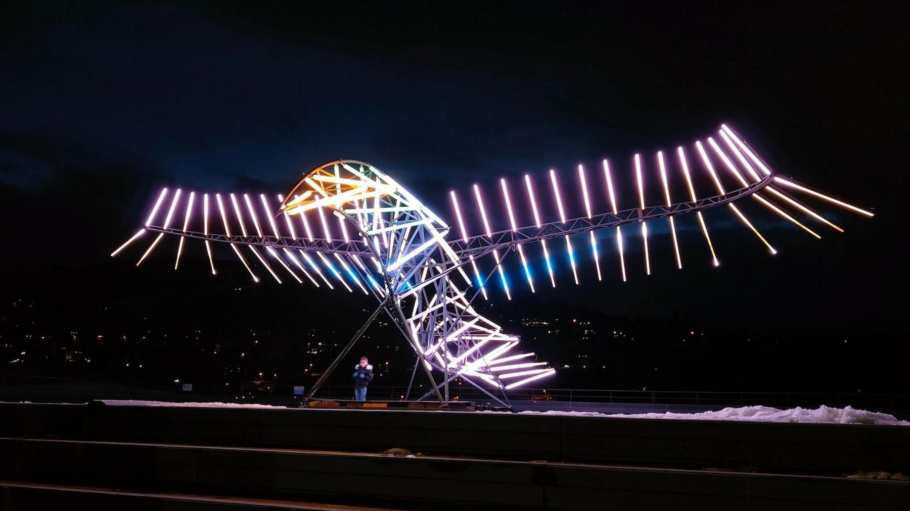 A large sculpture of a bird is stood looking towards the left of the picture. Its structure and wings are made up of several sticks lights of various colours including white, pink, blue and yellow.