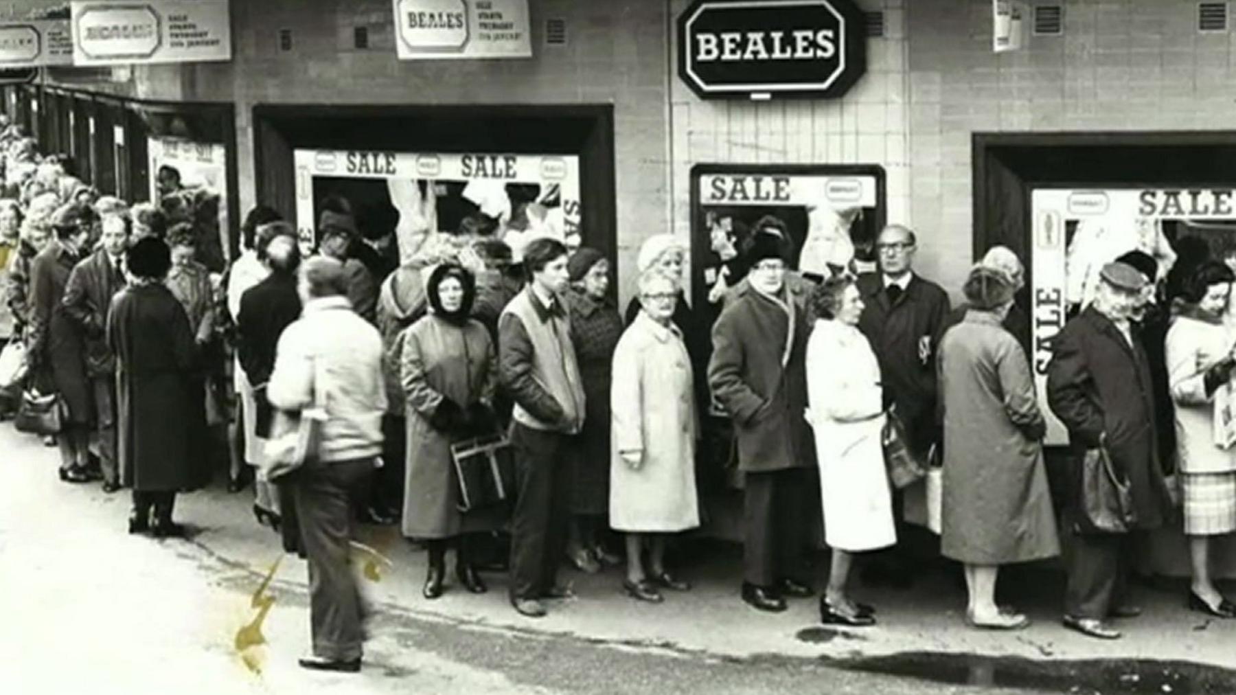 A black and white image of a large queue outside Beales with people dressed in 1960s-era clothing