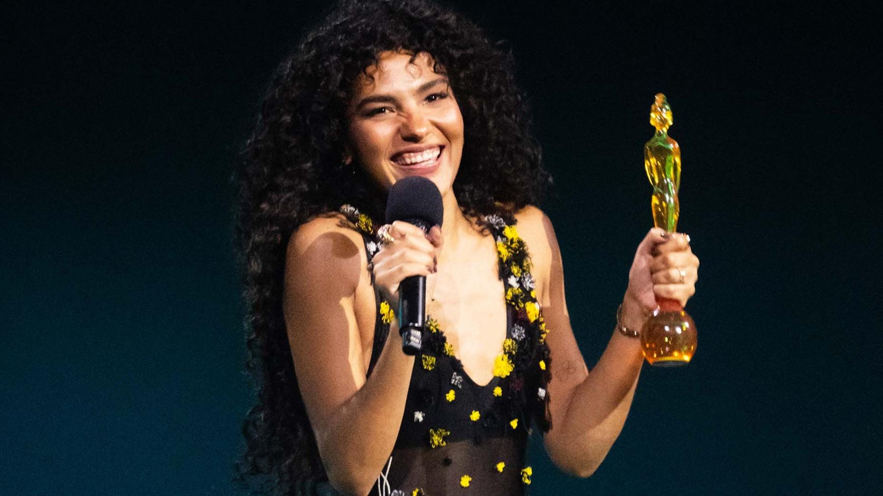 Olivia Dean smiling holding a brit award trophy  at the 2026 Brit Awards. She is wearing a black dress with yellow spots
