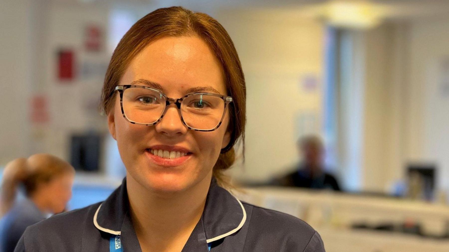 Sarah Rawsthorn, who is wearing blue scrubs and black-rimmed glasses, smiles in a reception area.